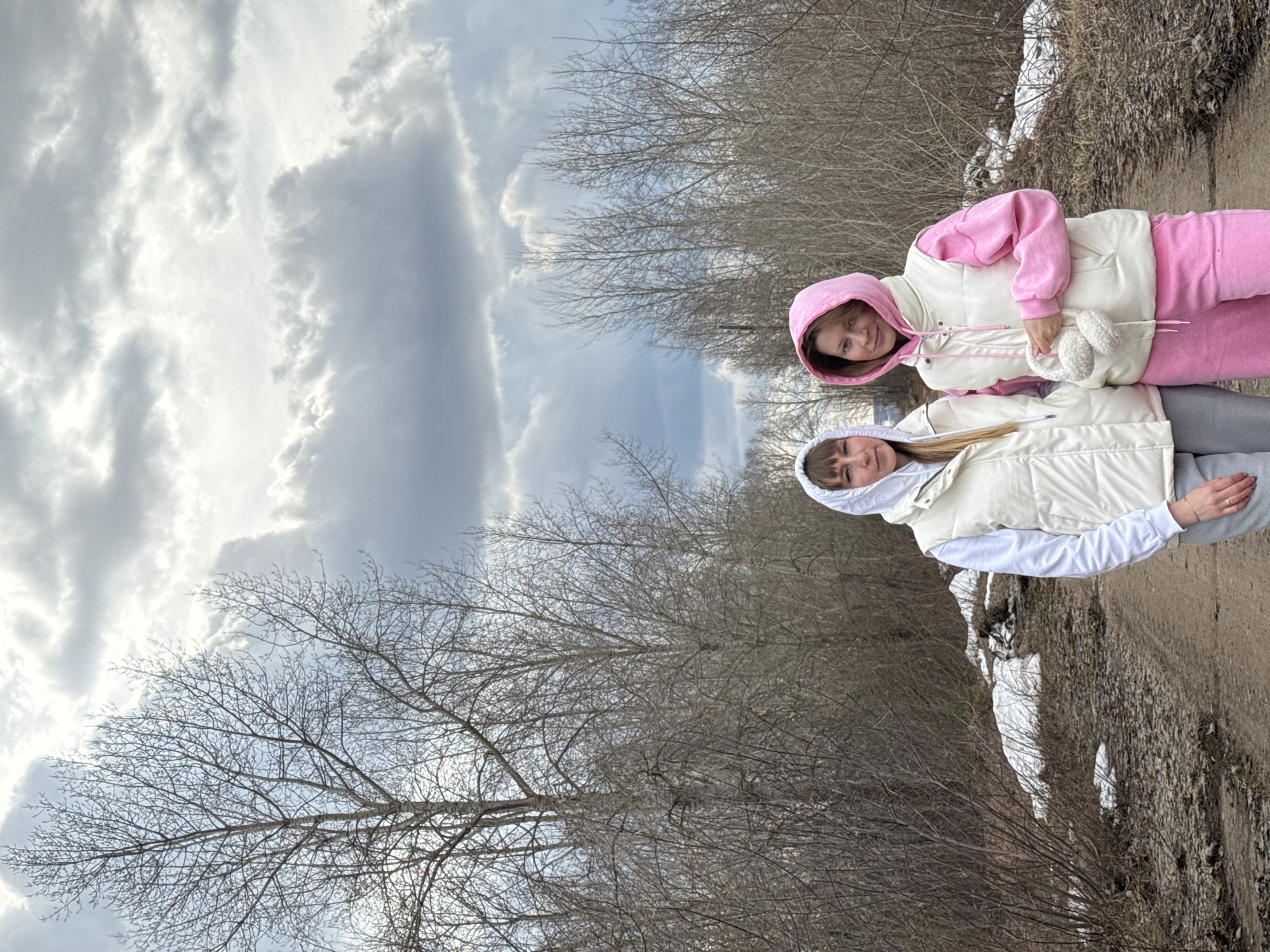 Two women are captured outdoors in what appears to be a late winter or early spring setting in Chaikovsky, Russia. The image is taken on an overcast day, with a cloudy sky dominating the upper portion of the frame. Bare trees, devoid of leaves, line the left side and background, indicating the cold season. Patches of snow are visible along a dirt path on the right side of the scene.
Both women are standing side-by-side, facing the camera with slight smiles, suggesting they are posing for a photograph. The woman on the left, who appears taller, has light-colored hair and is dressed in a white hooded sweatshirt, a light-colored (likely cream or off-white) puffy vest, and light grey sweatpants. The woman on the right, slightly shorter, has light brown or blonde hair and is wearing a pink hooded sweatshirt with a matching light-colored puffy vest and pink sweatpants. She is holding a pair of white fluffy earmuffs in her clasped hands in front of her. Their warm attire further emphasizes the chilly weather conditions. No text is visible in the image.