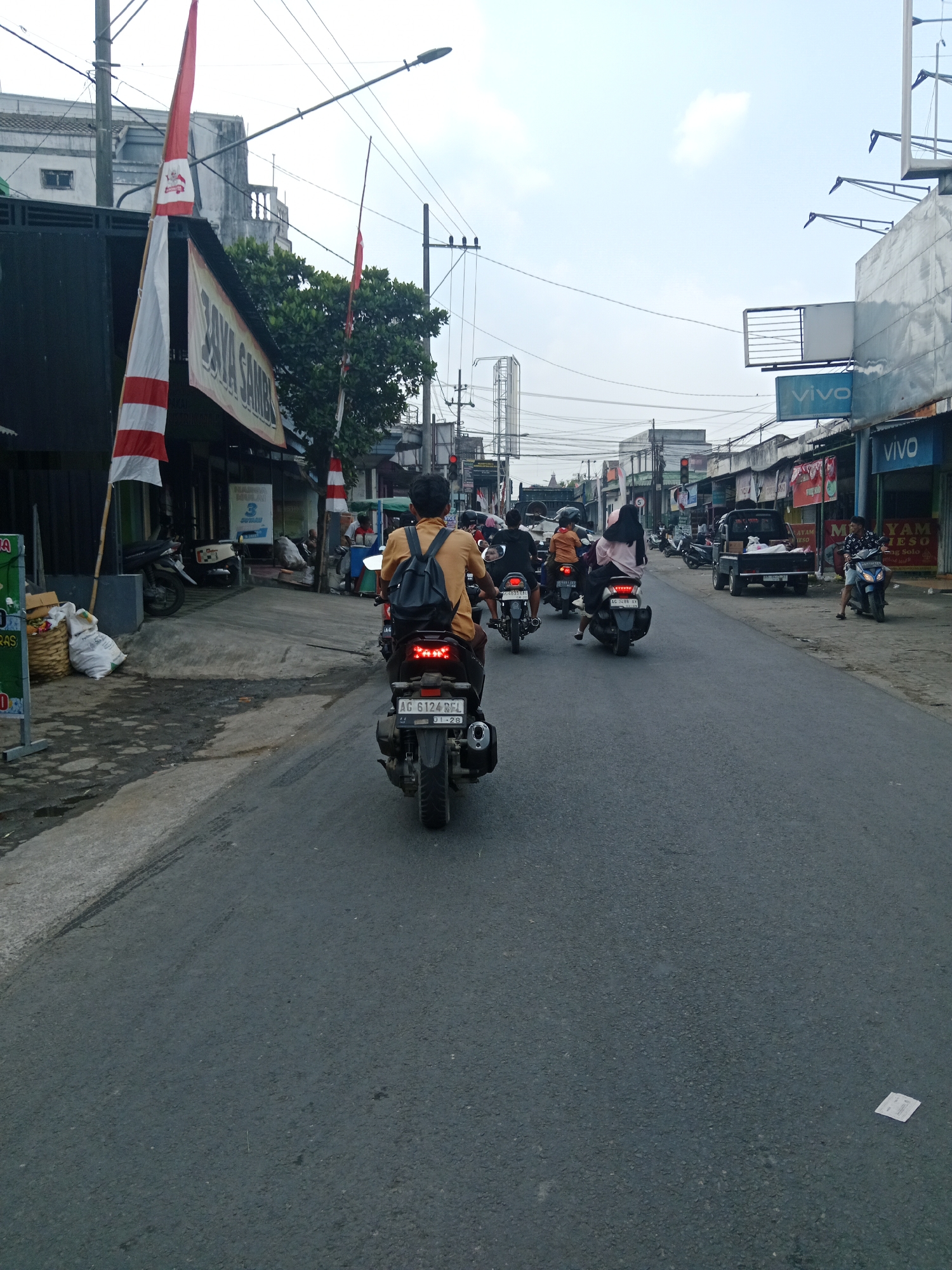 The image shows a street scene in Sambi, Indonesia, with several motorcycles and a small truck. In the foreground, a person wearing a yellow shirt and a backpack rides a motorcycle. The motorcycle has a license plate visible: "AG 6124 BGL". On the left side of the street, there are shops with banners and flags, including what appears to be an Indonesian flag. Several other motorcycles are seen on the road, some with passengers. In the background, buildings and overhead power lines are visible, indicating an urban or semi-urban environment. The sky is overcast.
