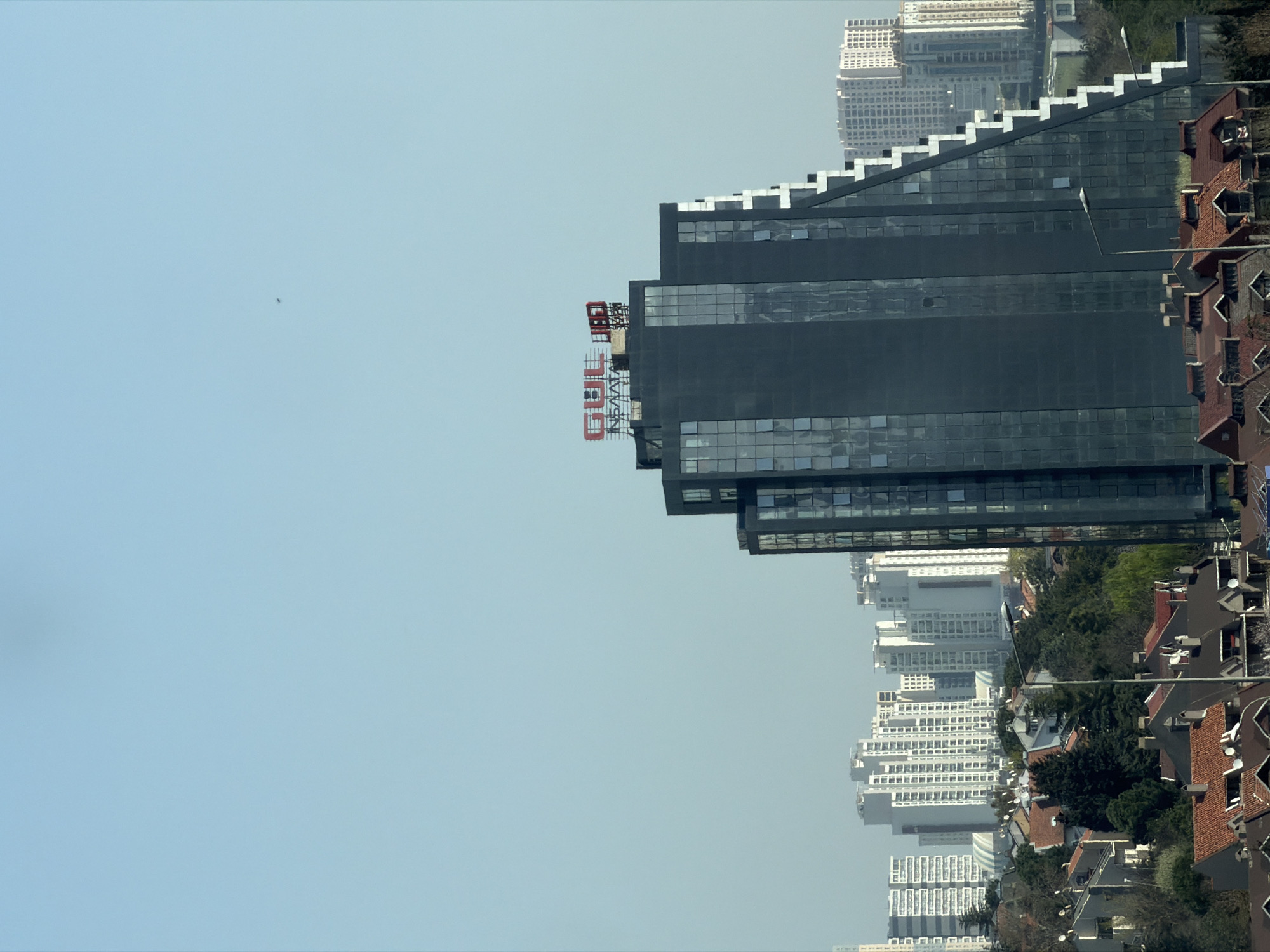 The image shows a cityscape, with the sky taking up the majority of the left side. On the right, a tall, dark building with stepped layers is prominently featured, bearing a red and white sign with the letters "GBIG" at the top. Below the building, there are other high-rise buildings and houses with red roofs, interspersed with trees. The buildings and houses suggest a dense urban environment. Based on the information provided, I can tell you that this is most likely an image of Istanbul and not Avcılar. This would be based upon the architecture and building structure of the photograph.