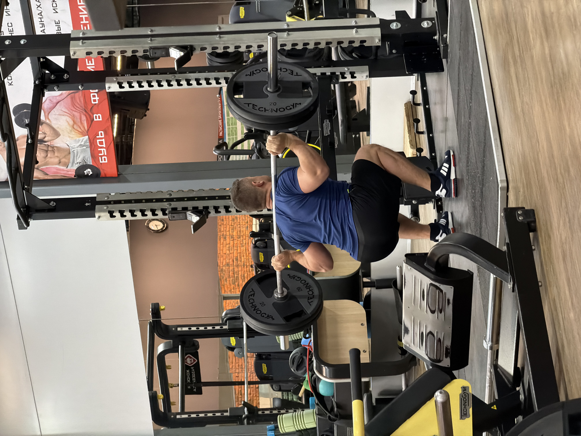 The image depicts an indoor scene within a modern gym or fitness center in Chaikovsky, Russia. The primary subject is a man, viewed from behind, performing a weightlifting exercise. He is wearing a blue short-sleeved t-shirt and black athletic shorts, along with athletic shoes featuring white stripes. The man is positioned beneath a barbell, which is loaded with weight plates and rests on his upper back/shoulders, indicating he is engaged in a squat or similar compound movement. He is currently in a deep squat position.
The equipment surrounding him includes a robust black squat rack or power cage, visible with numerous adjustment holes and safety pins. The barbell features "TECHNOGYM" branding on its black weight plates, which are marked with "20," likely indicating 20 kg. In the background, other gym equipment and machines are discernible. The floor consists of a dark, rubberized matting transitioning to a light wooden floor.
Visible text and branding in the gym include "TECHNOGYM" prominently on the weight plates and other machinery. On various posters and signs, primarily in Cyrillic (Russian), fragments of text can be seen: "Будь в форме" (Be in shape), "Расписание занятий" (Class schedule), and "ЖЕНСК" (likely part of "Женский," meaning Women's/Female). Other partial texts include "НЕС ИР," "АУНА/ХА," and "ВЫЕ ИСК." Numerical markings like "111111" are also visible on the squat rack's adjustment points. The overall atmosphere suggests a well-maintained and functional fitness environment.