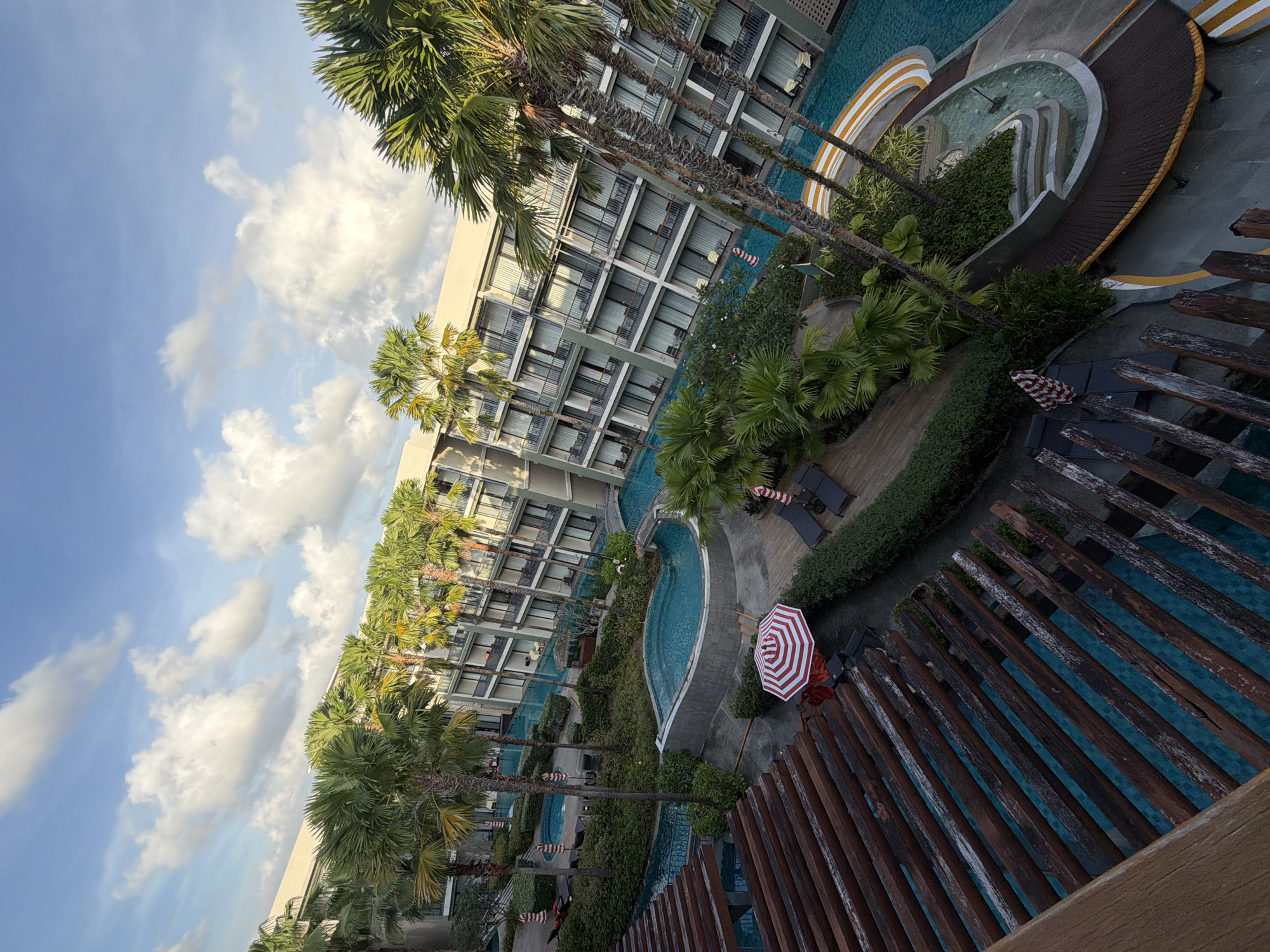 The image shows a resort swimming pool area at the Bang Muang Hotel in Bang Muang, Thailand. The swimming pool is a lagoon-style pool with turquoise water and a bridge crossing it in the distance. The pool is surrounded by lush green vegetation and palm trees. There are several lounge chairs and umbrellas around the pool. The hotel buildings are visible in the background, with balconies overlooking the pool. The sky is blue with white clouds. The image appears to have been taken from a high angle, possibly from a balcony or a higher floor of the hotel. The overall scene depicts a tropical resort paradise.