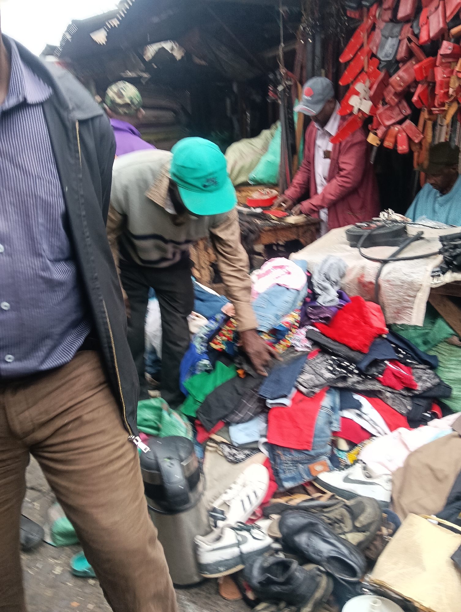 The image depicts a busy street market scene in Nairobi, Kenya, possibly in a bustling area like Eastleigh or along Dubois Road, known for their informal trade. In the foreground, the left side of the frame is dominated by the midsection and legs of a person wearing a dark striped shirt and brown trousers, with a black jacket zipped up to their waist. Their right hand is visible, resting near a pile of colorful clothing.
To the right, the scene unfolds with several individuals engaged in market activities. A person wearing a light gray long-sleeved shirt and a green cap is bent over, appearing to examine or sort through a large pile of clothing and footwear. Adjacent to them, a person in a red jacket and a light-colored shirt is standing behind a stall, with their hands actively working with small items, possibly related to leather goods or accessories displayed on the wall behind them – a collection of red leather pouches or cases are prominently visible. Another individual, wearing a light blue patterned shirt, is seated further to the right, seemingly observing or interacting with the stall owner.
The ground is covered with a mix of debris, discarded items, and more merchandise, including shoes and various articles of clothing scattered around. The stalls themselves appear to be makeshift structures, with corrugated iron roofing and wooden elements, typical of informal market setups. The overall atmosphere is one of active commerce and a lively, albeit somewhat chaotic, urban environment.