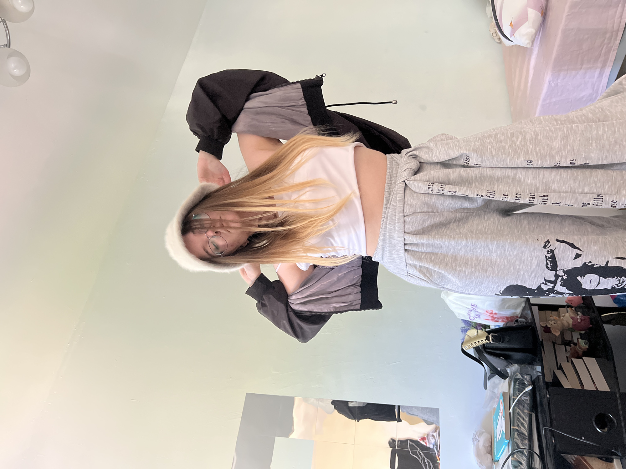 A young person with long blonde hair wearing glasses and a white fuzzy hat is looking directly at the camera. They are wearing a white crop top and gray sweatpants with writing on them. They are also wearing a black jacket with gray sleeves. The background consists of a light green wall, a bed with white sheets, a black table, and a reflective surface. The location appears to be indoors, potentially in a bedroom. The image was taken in Pushchino, Russia.