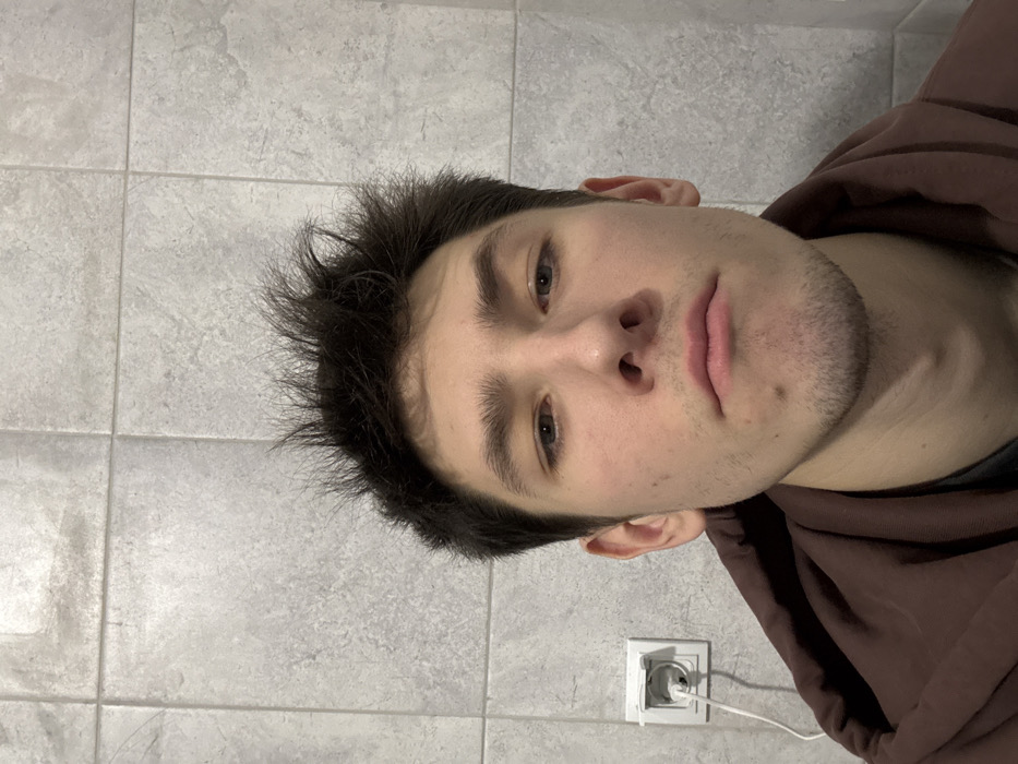 A young adult male is featured in a close-up, frontal portrait, taken indoors in Yanino-1, Russia. He has dark, spiked hair, light eyes, and minimal stubble on his chin. He wears a dark brown hooded garment over a darker shirt. The subject looks directly at the camera with a neutral expression. The background consists of light gray, rectangular wall tiles. To the left of the subject, a white electrical outlet is mounted on the wall, with a white cable plugged into its upper receptacle.