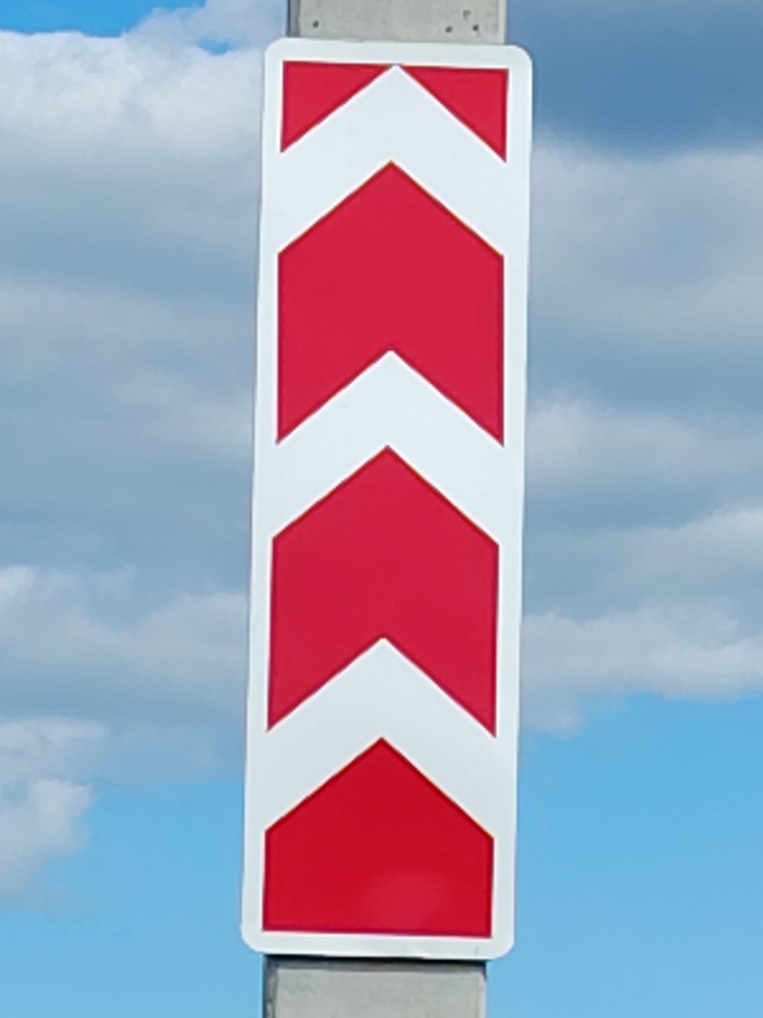 A vertical road sign with alternating red and white chevron shapes is mounted on a concrete post. The sign is positioned against a bright blue sky with scattered white clouds. The chevron shapes point downwards, with the white chevrons forming angles within the red rectangles. The sign appears to be a warning or directional marker for a curve in the road. The image does not contain any people or visible city features beyond the road sign itself. The location of Хоругвино, Russia, is mentioned, but the image itself does not provide specific visual cues to confirm this location.