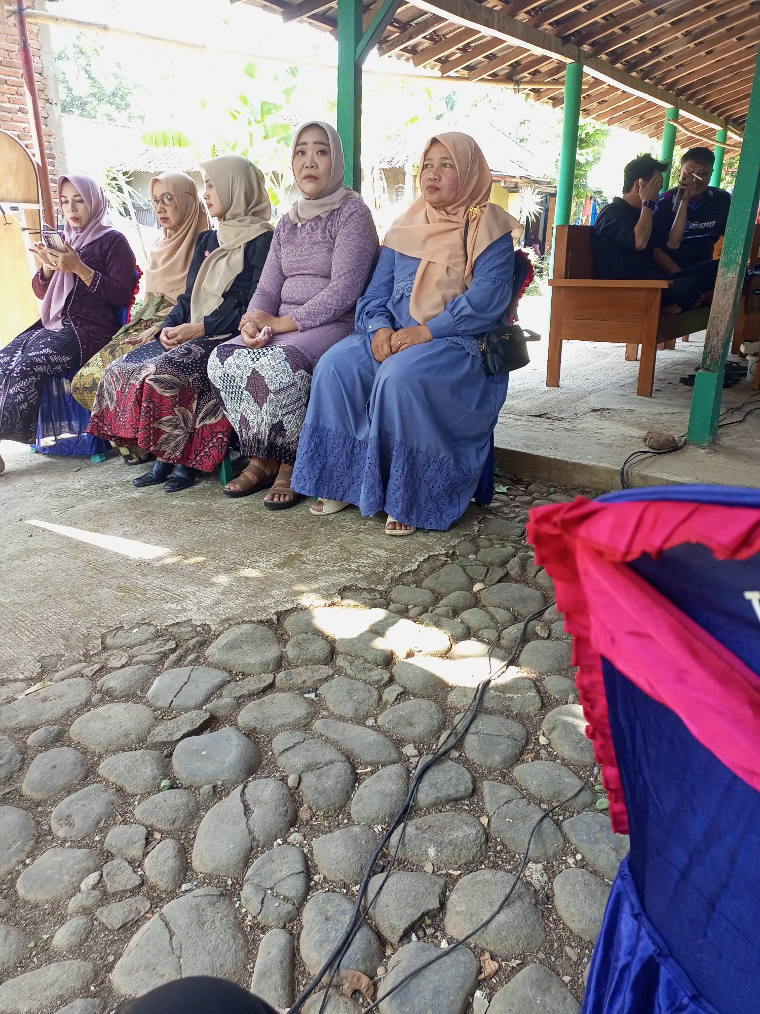 A group of five Indonesian women wearing headscarves and modest attire are seated on a concrete surface under a wooden shelter. The woman on the far left wears a dark purple top and a patterned skirt, and is looking at her phone. The next woman wears a light brown headscarf and glasses, with a patterned skirt. The third woman is wearing a beige headscarf and a dark top with a patterned skirt. The fourth woman wears a lilac lace top and a patterned skirt. The woman on the far right wears a light brown headscarf and a blue dress. Behind the seated women, two men are visible. One man in a black shirt is holding his face, and the other man in a blue shirt is smoking. The shelter has a wooden roof and green posts. The ground in front of the women is paved with large, irregular grey stones. In the foreground, a portion of a blue and pink draped fabric is visible. The background shows foliage and a brick wall. The scene appears to be outdoors in a public or community space, possibly at an event.