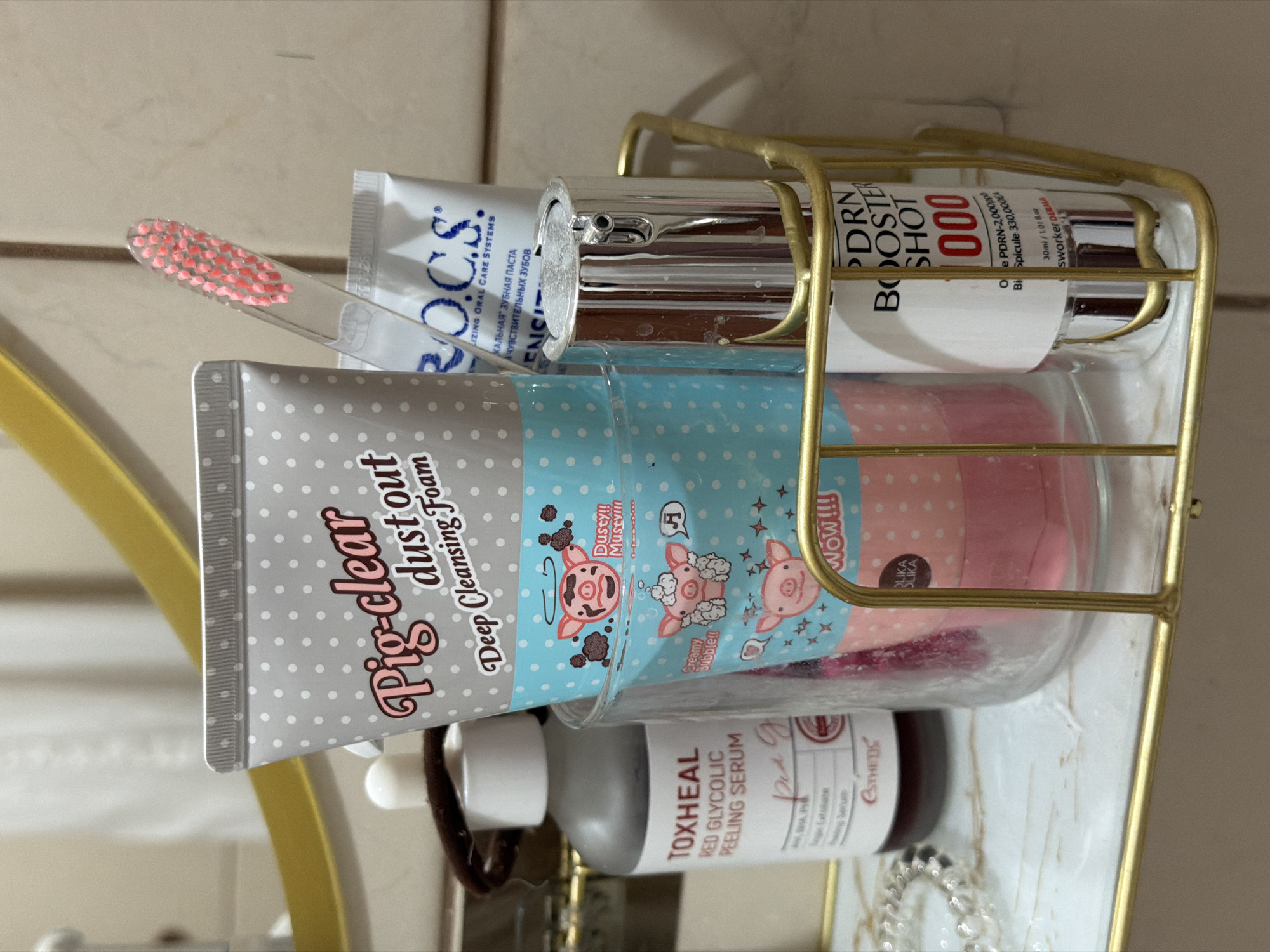 The image depicts a close-up, eye-level view of a bathroom shelf in Moscow, Russia. The shelf is made of white marble and has a gold wire organizer holding various cosmetic and personal hygiene items. In the center, a clear glass holds a pink cleansing foam tube decorated with cartoon pigs. To the left of the glass, a grey bottle labeled "TOXHEAL RED GLYCOLIC PEELING SERUM" is visible. To the right of the glass, a silver pump bottle labeled "PDRN BOOSTER SHOT 000" is present. A clear toothbrush with pink bristles rests on top of the cleansing foam tube. Behind these items, a white tube with blue lettering, "R.O.C.S.", is partially obscured. The background consists of a tiled wall and a portion of a circular gold mirror frame. The lighting suggests an indoor setting, possibly a bathroom. No individuals are present in the scene.