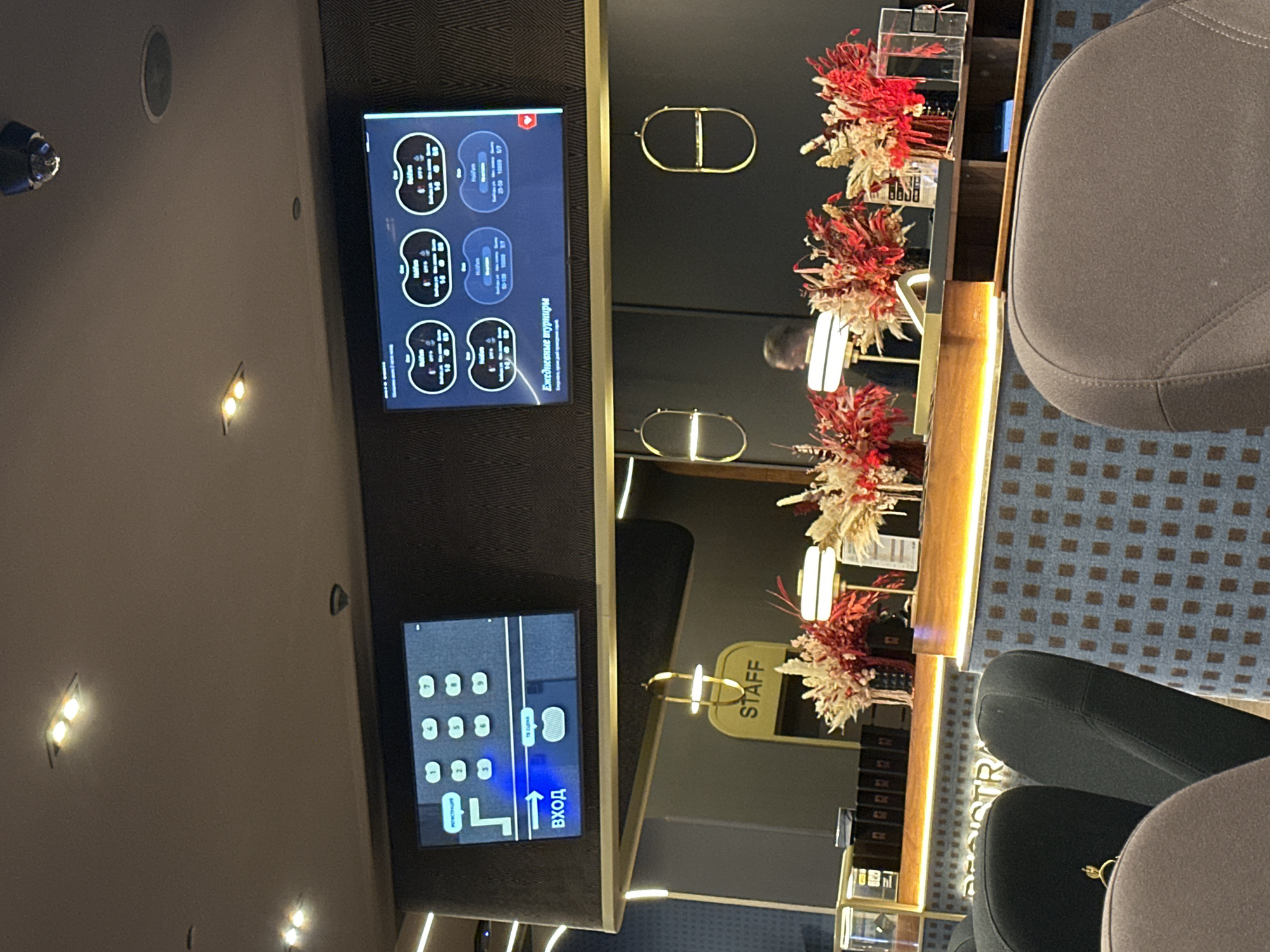 The image depicts an indoor scene, likely a registration or reception area. Two screens are visible. The left screen displays a digital keypad with numbers 1 through 9, alongside directional arrows and the Russian word "ВХОД" (Entrance). It also shows the word "РЕГИСТРАЦИЯ" (Registration) in the upper left corner, indicating the purpose of the screen. The right screen shows multiple circular displays, each with text and numbers, suggesting data related to games or tournaments, with the Russian text "Ежедневные турниры" (Daily Tournaments) below.
A prominent yellow sign with the word "STAFF" is hanging, indicating an area designated for employees. In the foreground, several chairs are visible, some are dark gray and appear to be part of a seating arrangement. The background features a counter decorated with floral arrangements, primarily in shades of red and beige. The overall environment suggests a modern establishment, possibly a casino, hotel, or entertainment venue. The city of Estosadok, Russia is mentioned as the location, which is known for its resort areas.