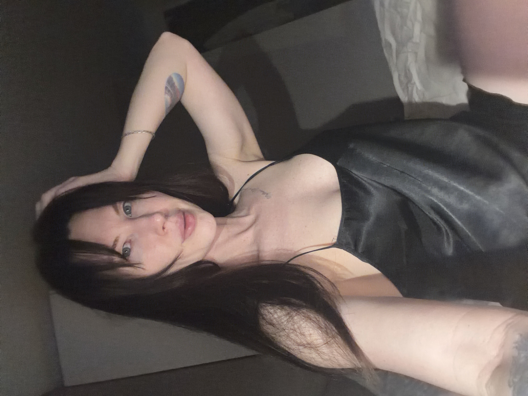 A woman with dark hair is lying down, her head tilted towards the camera. She is wearing a black sleeveless top and has a tattoo on her left arm. The background is dark and indistinct. There is no evidence to suggest the location is Perm, Russia.