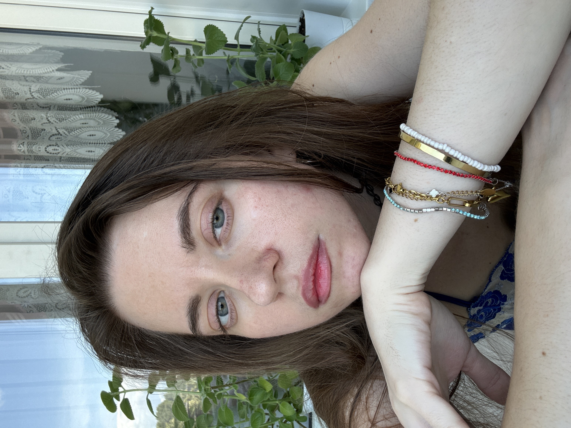 A woman with brown hair and blue eyes is leaning on her arm, resting her chin on her hand. She is wearing a blue and white floral top and several bracelets on her wrist. The background appears to be an indoor setting with a window and plants.