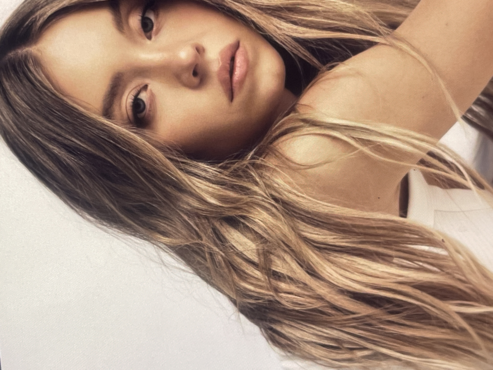 This medium close-up photograph captures a young woman in a reclining pose, oriented diagonally across the frame. She gazes directly at the viewer with a calm, composed expression and slightly parted lips. Her long, wavy hair, a blend of blonde and light brown hues, cascades around her face and over her right shoulder. A clear part is visible on her scalp. Her eyes are light, and her eyebrows are distinct. Soft, diffused lighting illuminates her face, creating gentle shadows that add depth to her features. A glimpse of a light-colored garment, possibly white, is visible near her shoulder. The background is a plain, off-white surface, suggesting an indoor studio setting or a simple backdrop. The image focuses intently on her face and hair, conveying a serene yet captivating presence. No specific time of day or weather can be determined from the image. There is no visible text. The photograph was taken in Poltava, Ukraine.