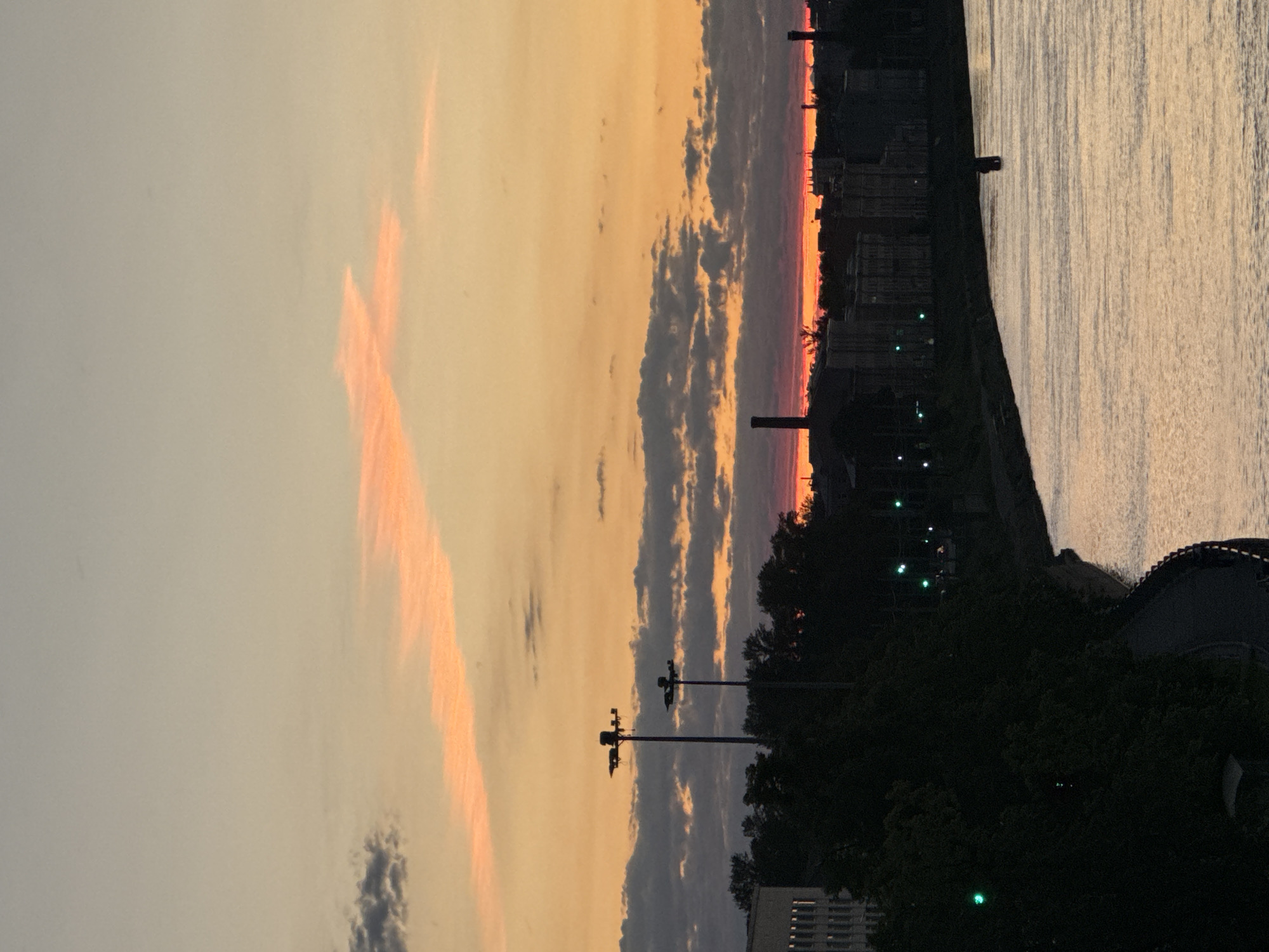 This image depicts a sunset over the city of Ladozhskii Trudposelok, Russia, with a river in the foreground. The sky is painted with hues of orange, pink, and pale yellow, with streaks of clouds running horizontally. In the distance, a silhouette of buildings and industrial chimneys can be seen against the colorful sky. A river reflects the sunset colors, with a small dark object, possibly a buoy or a submerged structure, visible in the water. The left side of the image shows a dark, tree-lined embankment leading to a pathway. Tall lampposts stand silhouetted against the sky. Green lights are visible along the embankment, suggesting streetlights or traffic signals. The overall scene is serene and atmospheric, capturing the quiet beauty of an evening in the city.