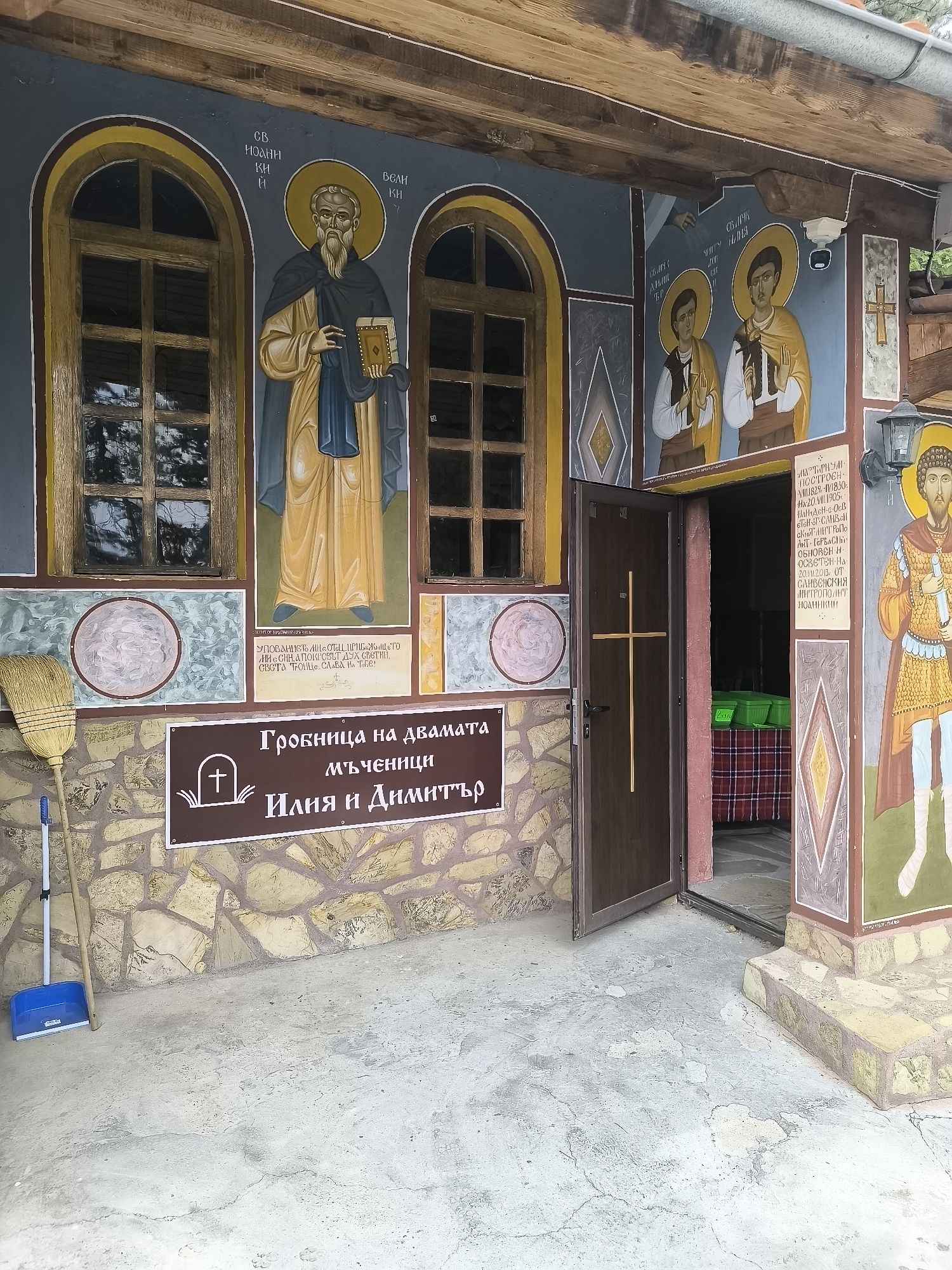 The image shows the exterior of a building, likely a chapel or church, in Chintulovo, Bulgaria. The facade is decorated with religious frescoes depicting saints. To the left, a broom and dustpan are leaning against the wall. A dark brown sign with white lettering, featuring a tombstone icon, is mounted on the wall above the broom and dustpan. The sign reads "Гробница на двамата мъченици Илия и Димитър" which translates to "Tomb of the two martyrs Iliya and Dimitar." The building has a wooden roof overhang supported by visible beams. An open wooden door with a vertical cross design in the center reveals a glimpse of the interior, which appears to have a red and white checkered cloth and green containers. A lamp is mounted on the right side of the building next to a fresco of a saint in armor. The ground in front of the building is a concrete surface with some visible cracks.