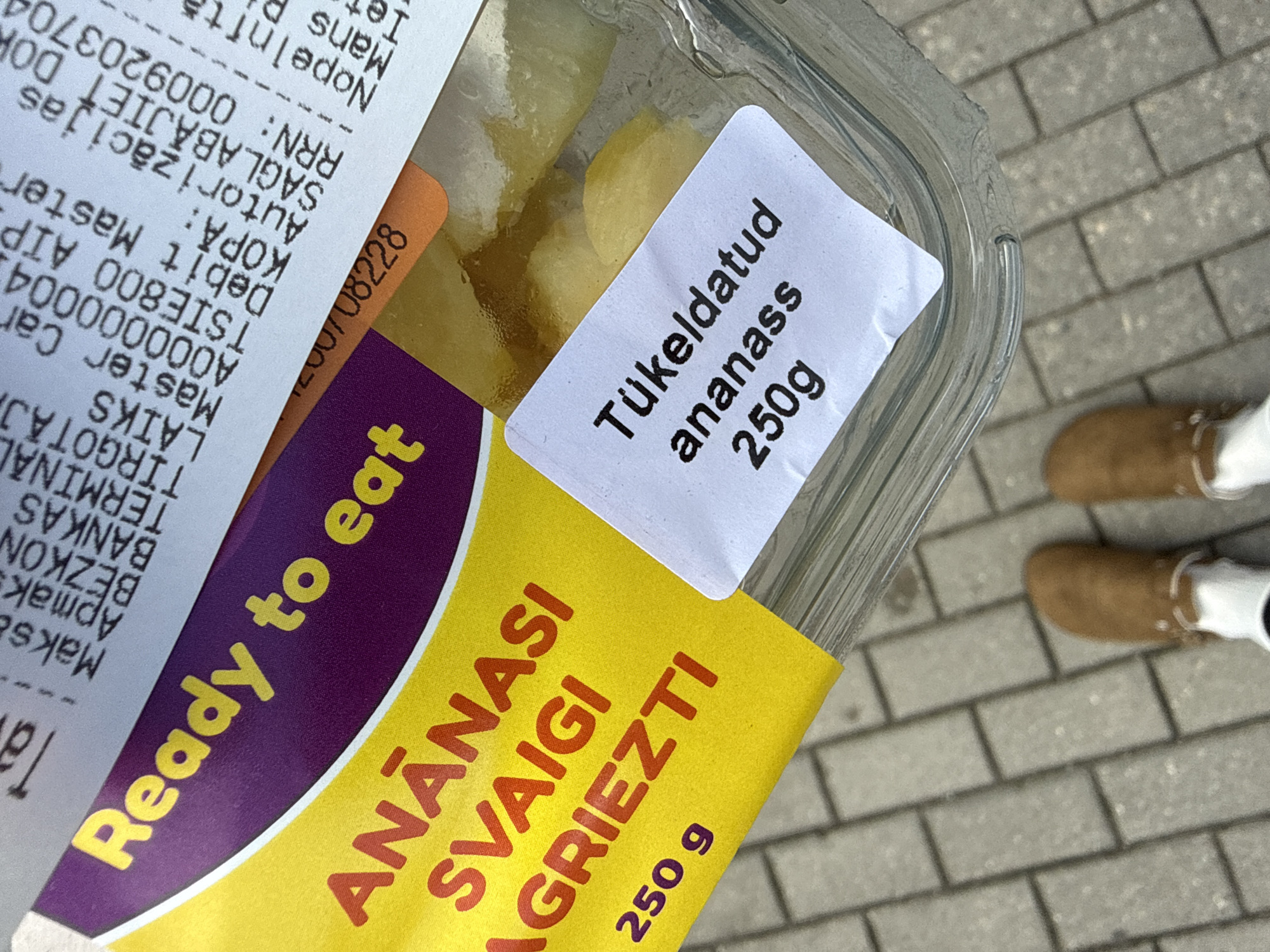 The image shows a close-up of a clear plastic container of pre-cut pineapple, likely purchased in Riga, Latvia, as evidenced by the Latvian text on the receipt and packaging. The container is placed on a grey paved sidewalk. The packaging has a yellow sticker with red text that reads "ANANASI SVAIGI AGRIEZTI 250 g" and a purple band with the words "Ready to eat" in yellow. A small white label on the container reads "Tükeldud ananass 250g". A portion of a crumpled receipt is visible, showing transaction details with Latvian text such as "Maks.", "BEZKON", "BANKAS", "TERMINAL", "LAIKS", "Master Car", "TSİE800 AİP", "KOPA:", "Debit Master", "Autorizācijas", "SAGLABĀJIET DOK", and "RRn: 0009203704". The bottom of the image shows a person's feet wearing brown boots and white socks, standing on the sidewalk. The scene appears to be an outdoor setting in a city environment.