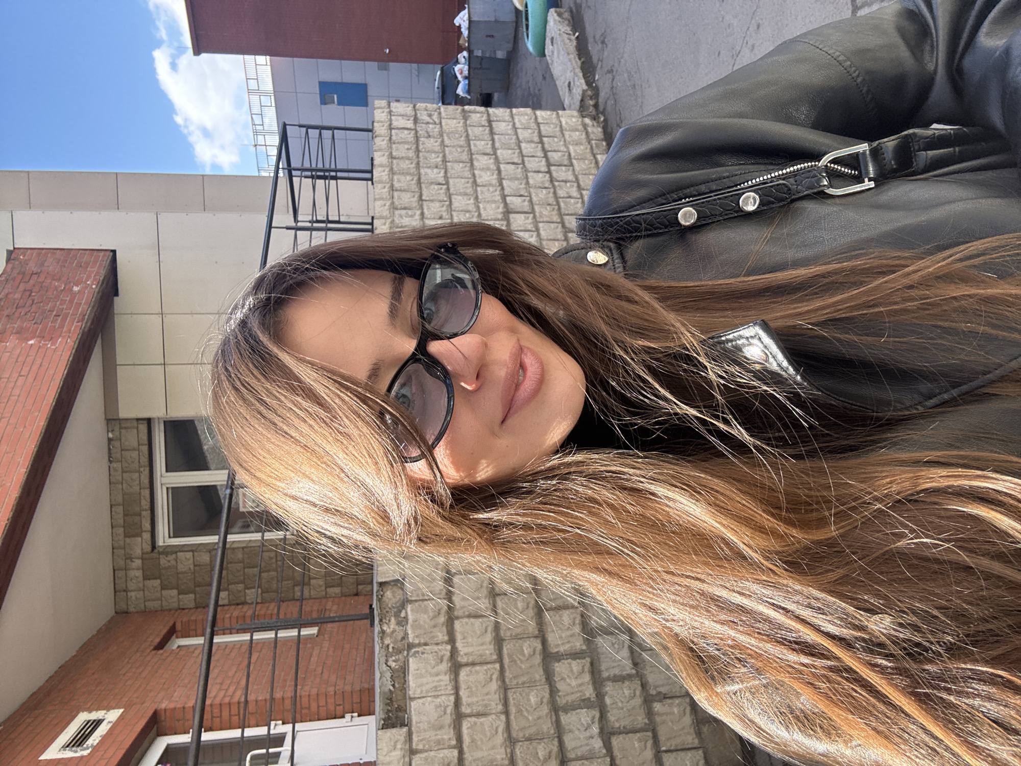 A woman wearing glasses and a black leather jacket stands outside in what appears to be Krasnoyarsk, Russia. Buildings with brick and concrete facades are visible in the background. The sky is partly cloudy.