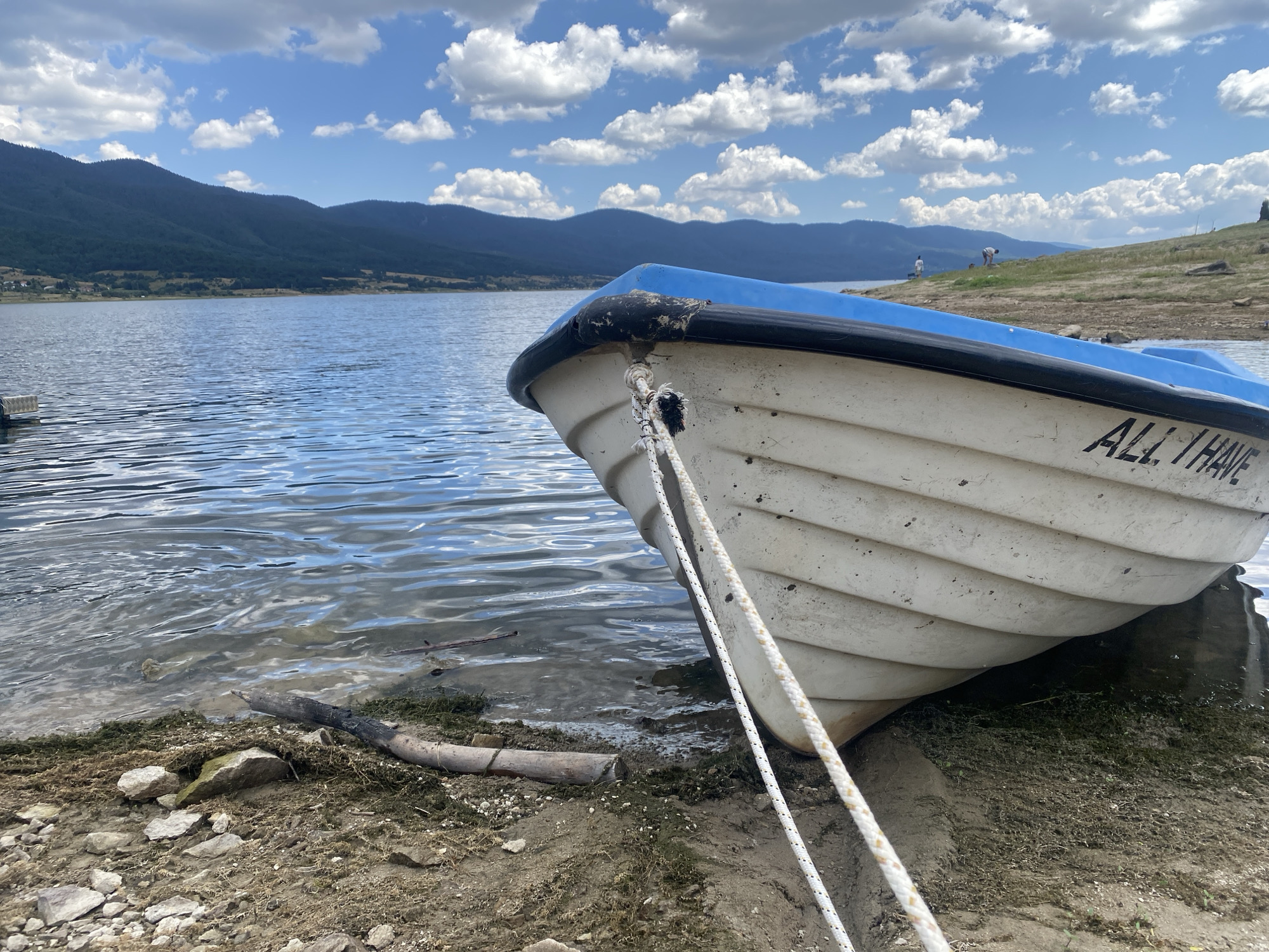 In the city of Sarnitsa, Bulgaria, a white boat with a blue rim is pulled ashore on a dirt and gravel bank next to a calm body of water. The boat is partially submerged, with its bow facing towards the camera. A white rope is tied to the bow and extends diagonally across the image. On the side of the boat, the words "ALL I HAVE" are visible in black lettering. In the background, a large body of water stretches out towards a line of dark green, tree-covered mountains under a sky filled with fluffy white clouds. A few small figures are visible on the grassy hillside in the distance. The scene appears to be a peaceful, natural landscape.