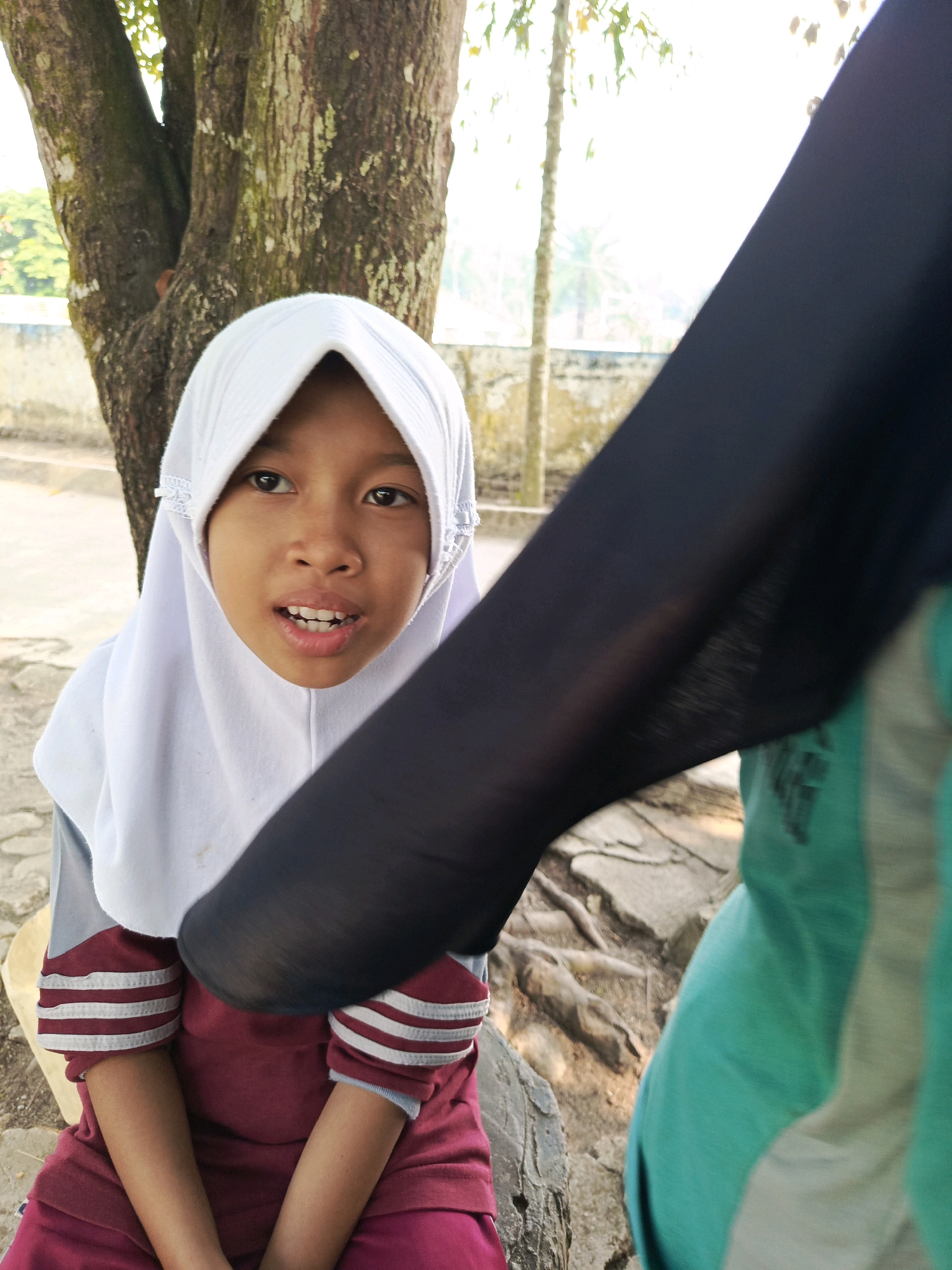A young girl wearing a white hijab and a maroon shirt with white stripes on the sleeves is seated outdoors near a tree. Her mouth is slightly open, revealing her teeth. A person wearing a black garment and a green top is partially visible in the foreground, obscuring part of the girl's right side. The background features a large tree trunk, exposed tree roots on the ground, and a wall with foliage visible beyond it, suggesting an outdoor setting in Purwakarya, Indonesia. The lighting indicates it is daytime.