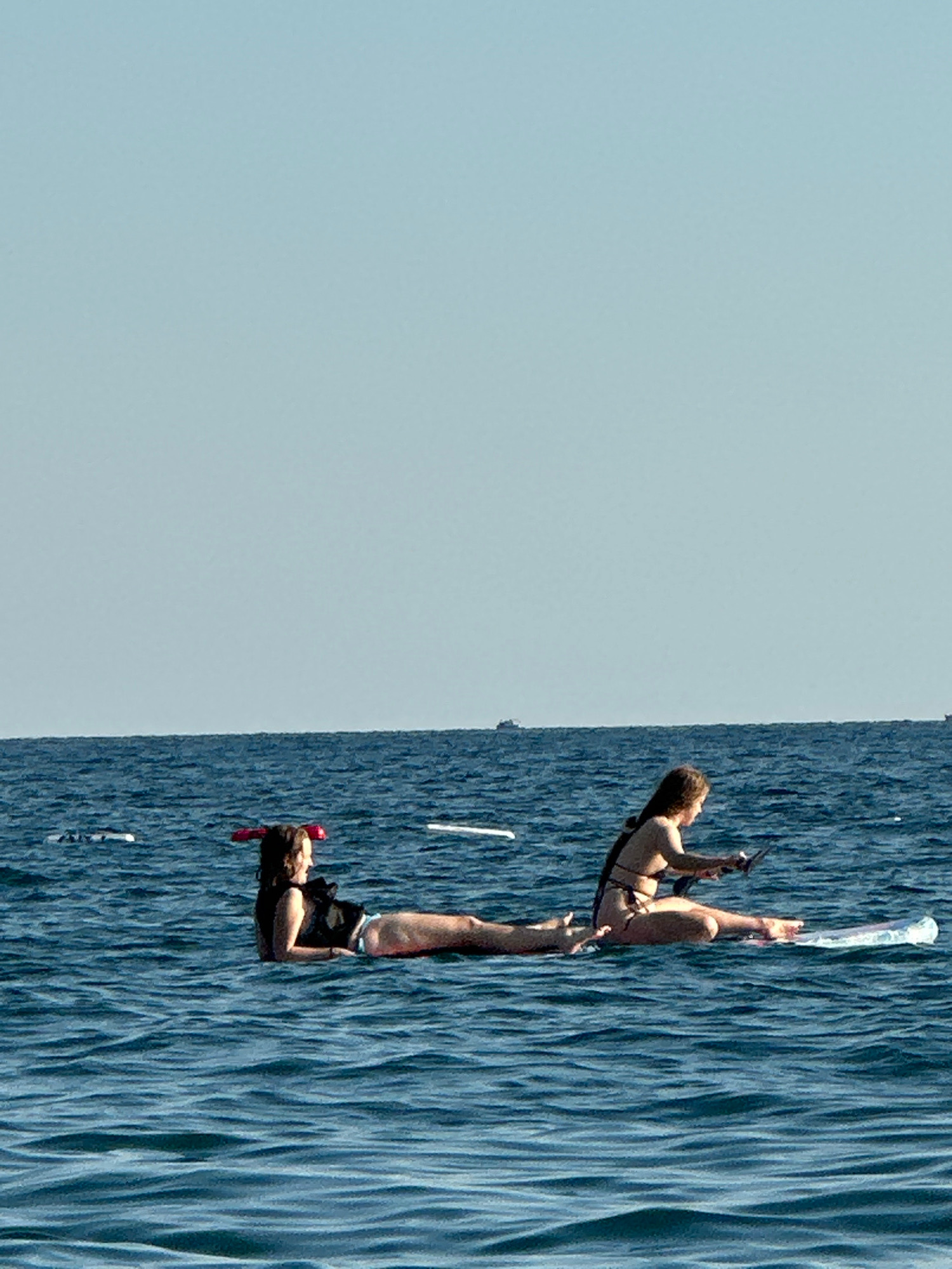 Two individuals are on paddleboards in the Adriatic Sea near Boreti, Montenegro. The person on the left, wearing a black top and blue bottoms, lies on their stomach with a red snorkel mask on their head. The person on the right, wearing a bikini, is seated on their paddleboard, looking down at a phone. Both paddleboards are white and floating on calm, blue water under a clear sky. In the distance, several boats are visible on the horizon.