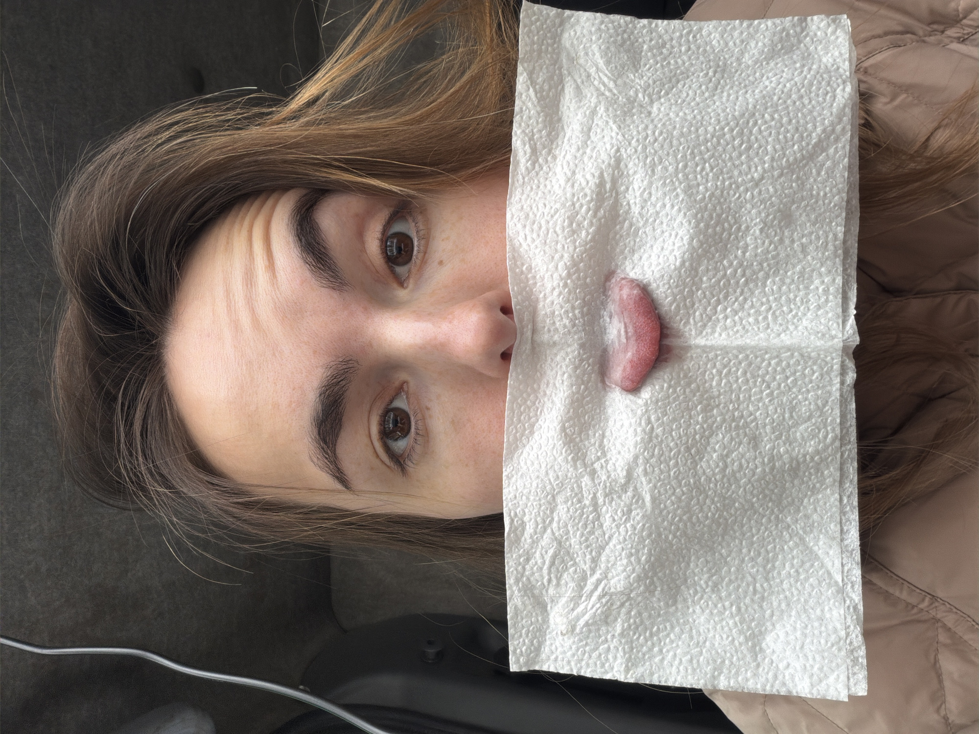 A woman with light brown hair and light skin is holding a white paper towel with a pink, fleshy mass on it in front of her face. The background shows a dark grey surface to her left and a light brown, quilted surface to her right. The scene does not provide information to determine its location within Izhevsk, Russia, or any location.