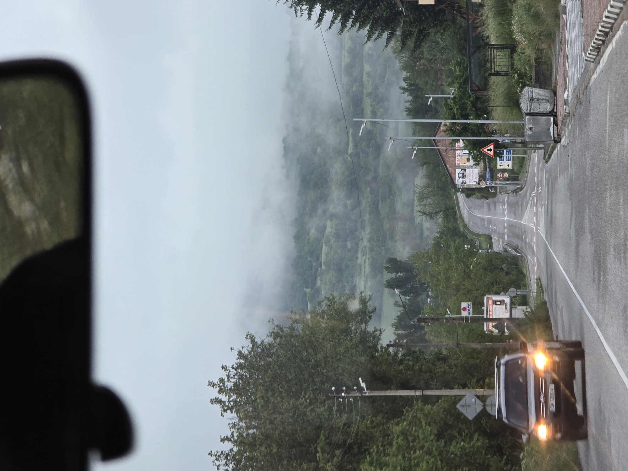 The image, captured from the perspective of a vehicle's interior (evidenced by a blurred portion of a mirror or frame on the left), depicts a rural road scene on a misty or foggy day. A two-lane asphalt road, wet from what appears to be rain or heavy condensation, curves gently to the right and recedes into the distance. A dark-colored car with its headlights illuminated is approaching from the foreground, partially visible on the right side of the road.
The landscape is characterized by dense green foliage and trees covering hillsides, which are largely obscured by thick mist or fog, especially in the background. Along the right side of the road, there is an intersection marked by several road signs. Visible text in Cyrillic script includes "РАДУШИ" (Radushi) and "ШОРКИ" on a signboard. Other signs include a red triangular warning sign with an exclamation mark, a blue rectangular directional sign with multiple arrows, and a sign indicating the end of a 25 km/h speed limit.
Further along the road on the right, a building with a red-tiled roof is visible amidst the trees, along with utility poles and wires. Two trash bins are situated beside a gate near the road. The overall ambiance is quiet and atmospheric, suggesting travel through a mountainous or hilly region under overcast conditions, likely in an Eastern European country given the Cyrillic script on the signs.