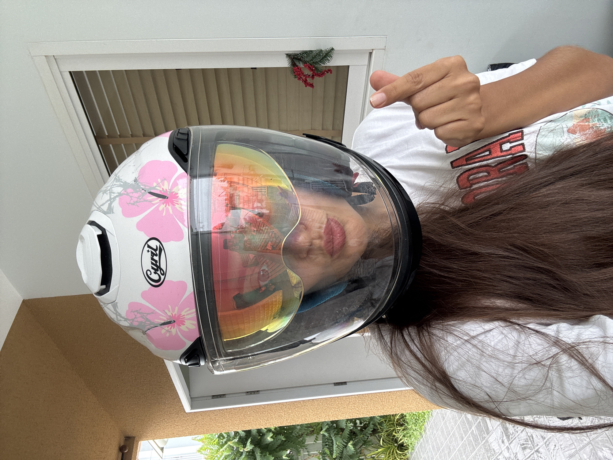 A young adult woman is positioned sideways, wearing a white motorcycle helmet adorned with pink floral designs. The helmet has a transparent visor, reflecting an interior scene. The woman is wearing a white t-shirt with text on the front. Her hair, which is dark brown, falls across the t-shirt. Her hand is in the upper right corner, making a hand gesture.
The background includes a window with white blinds. A small sprig of artificial pine needles and red berries is attached to the window frame. Outside, there are green plants, and the wall has a textured, light brown finish. This image was taken in Pa Khlok, Thailand.