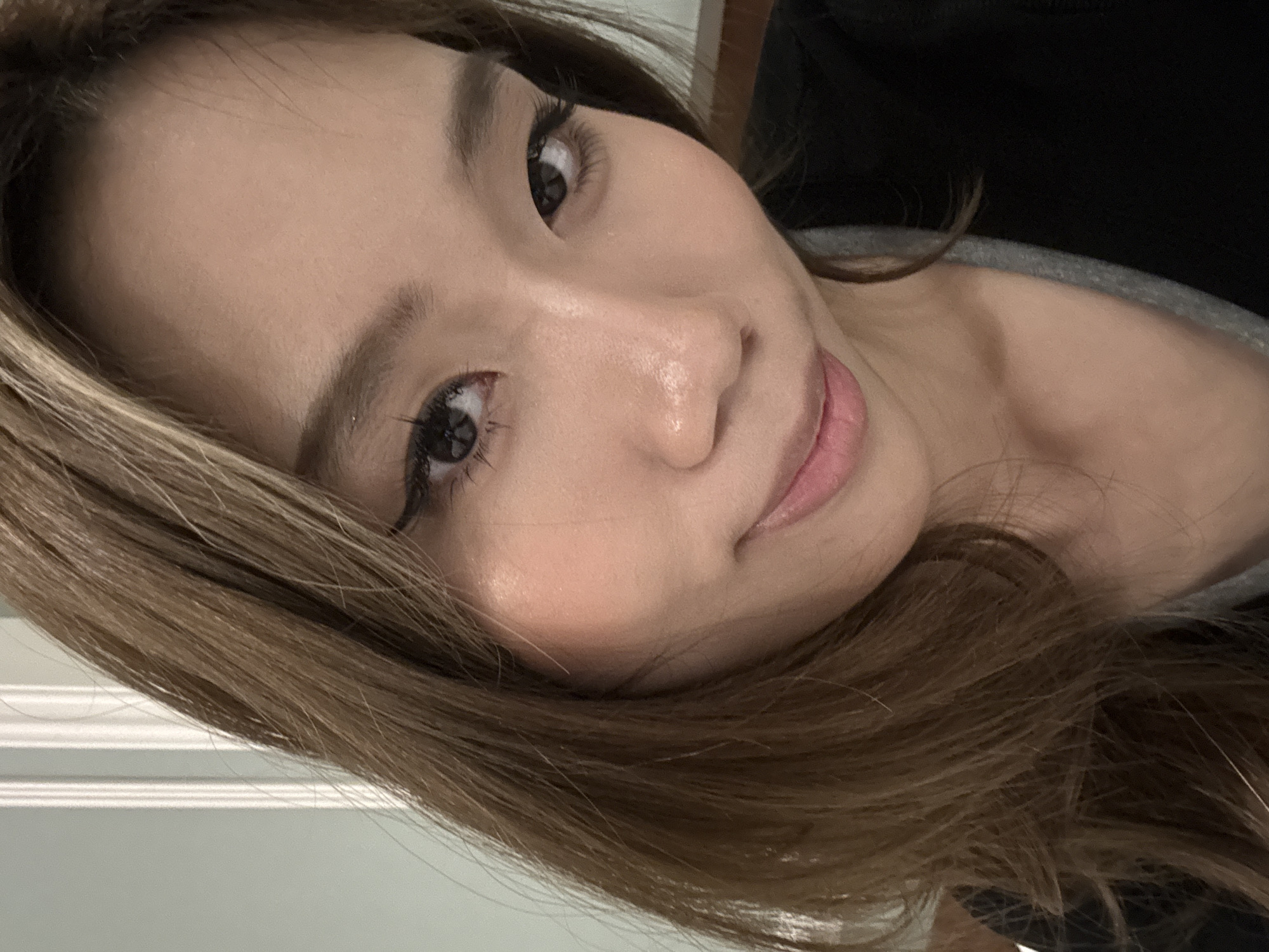 A close-up portrait of a woman with light brown hair, angled slightly to the left. She has dark eyes with eyeliner and long lashes. Her skin tone is fair with pink blush on her cheeks. She is wearing a gray shirt under a black top. The background appears to be a white wall with a decorative molding. There is no indication of this image being taken in Lanzhou, China.