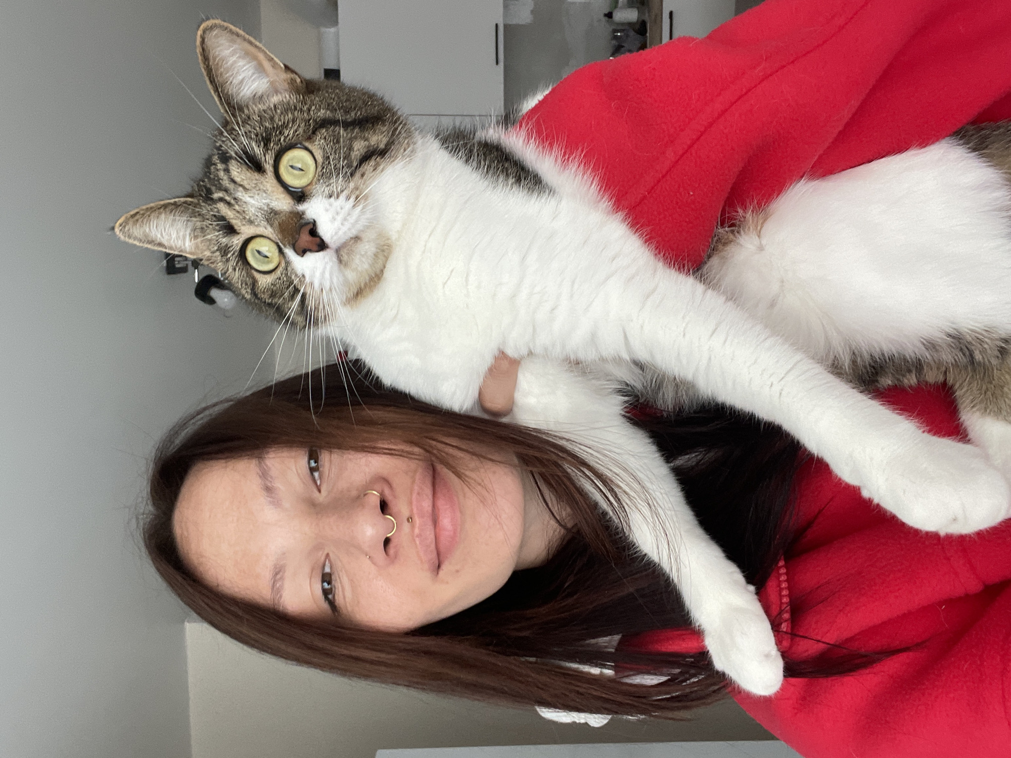 A woman is holding a cat. The cat is a tabby and white domestic shorthair. The woman has long brown hair and is wearing a red sweatshirt. She has piercings in her nose. The background is out of focus, showing part of a room with white walls and some cabinets. There is no indication of the location being Izhevsk, Russia.