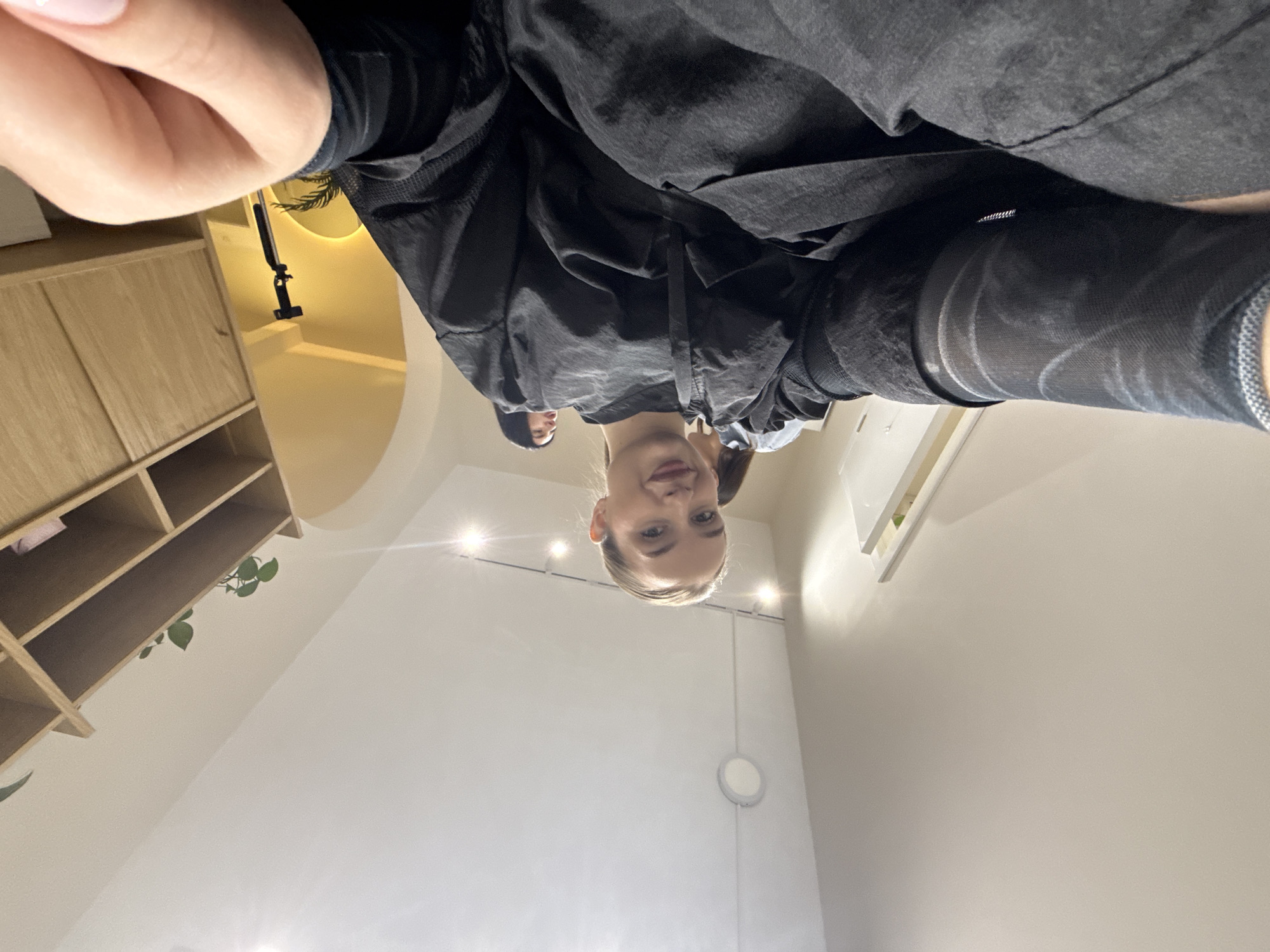 An indoor scene is captured from a very low-angle perspective, looking upwards. A young woman with blonde hair, pulled back, is the primary subject, positioned above the camera and looking down into it with a subtle, pleasant smile. She wears a dark, possibly black, long-sleeved top or jacket, and part of her light-skinned hand is visible in the immediate foreground. Another person with dark hair is partially visible behind her.
The background reveals a bright, modern interior. To the left, a light wooden shelving unit or cabinet with multiple open compartments is visible, displaying a small green plant. White walls with several bright, recessed spotlights in the ceiling and a circular white wall light are on the right. In the mid-ground, a black selfie stick or camera stand is visible against a yellow-lit decorative niche, which features the silhouette of a plant, possibly a palm frond. The overall setting is a clean, contemporary room, suggesting a salon, studio, or modern private space in Budva, Montenegro. There is no visible text.