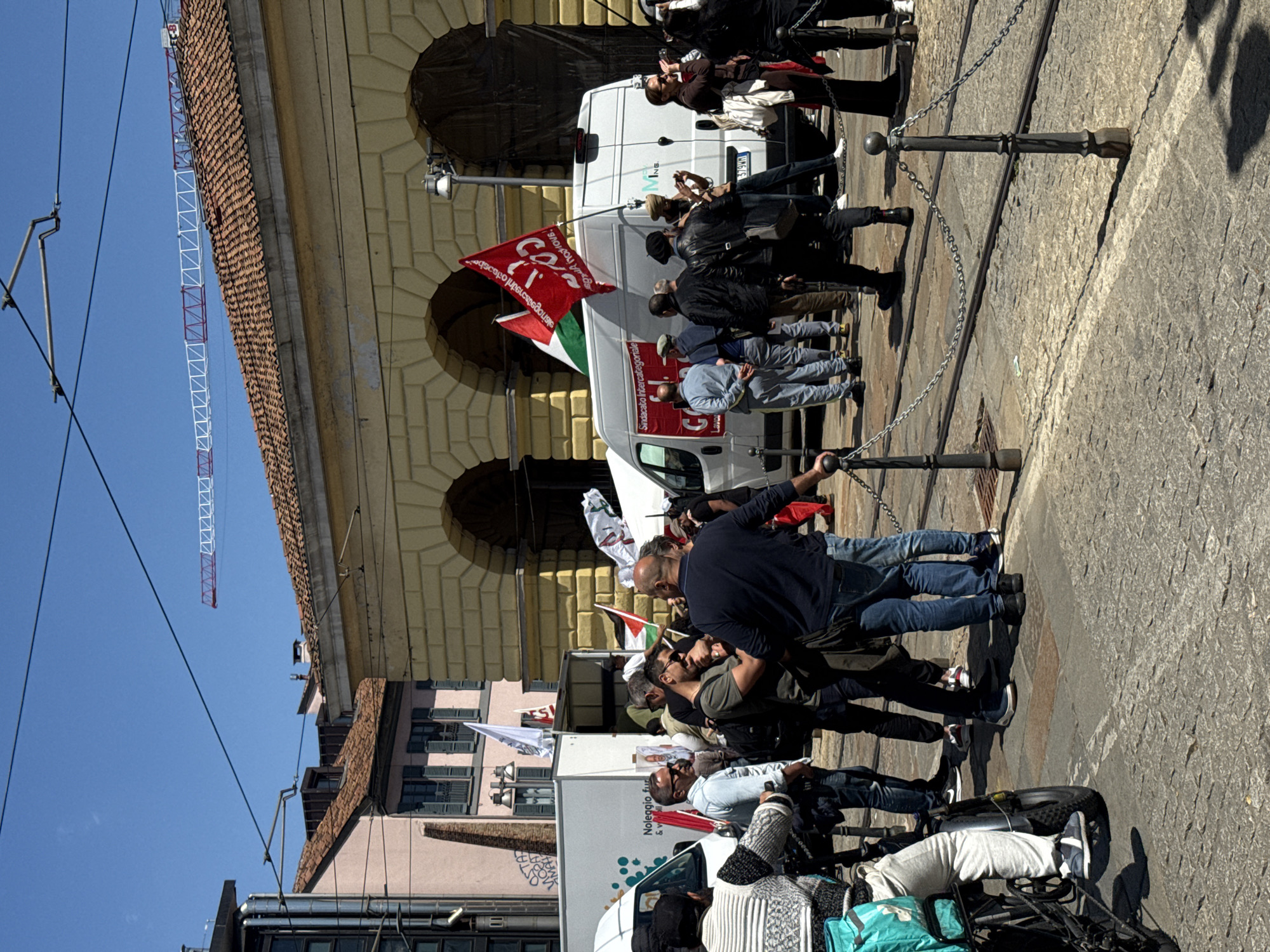 The image captures a demonstration in what appears to be a public space in Milan, Italy, likely a plaza or large pedestrian area. A crowd of people is gathered, some of them holding flags; one distinctly features the Italian flag. They are standing near a series of metal posts connected by chains, possibly forming a barrier. There are several white vans present; some have writing visible that includes “Noleggio fur” and “Sindacato Intercategoriale”. The architecture in the background includes a building with arched windows and a tiled roof. A construction crane is visible against a clear blue sky. Some individuals are holding phones and may be recording or taking pictures of the event.