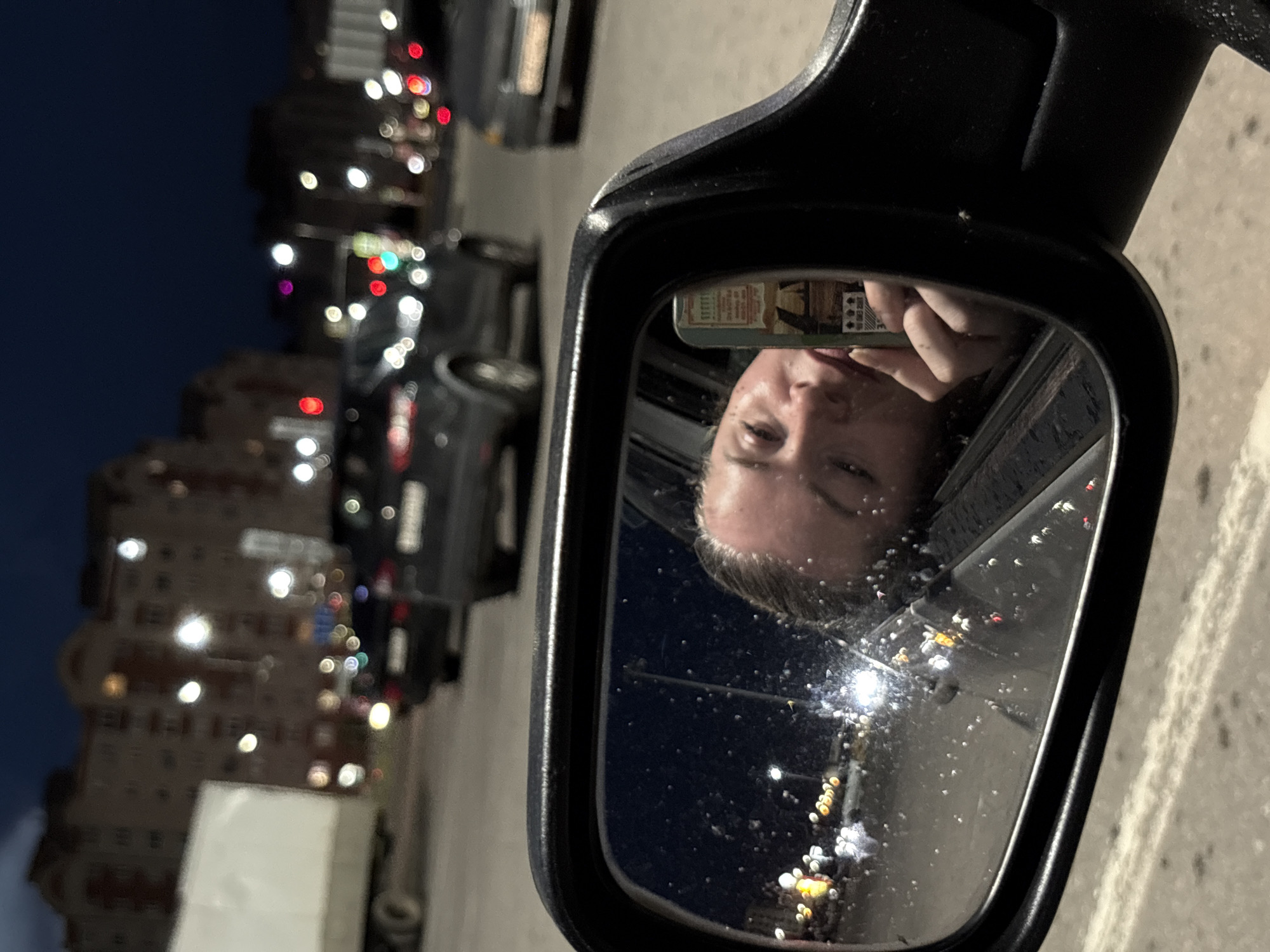 A person is taking a selfie in a car's side-view mirror at night, in the city of Veliky Novgorod, Russia. The person is leaning out of the car window, with their face filling a significant portion of the mirror's reflection. They are holding a mobile phone in their right hand, which is also reflected in the mirror, and appears to be taking the photo. The background reflected in the mirror shows streetlights and other vehicles on a road, indicating an urban environment. The car is likely in motion or stopped at a traffic light, given the blurry lights of the city in the background. The visible part of the car's exterior includes the frame of the side-view mirror. The overall scene suggests a casual moment captured while traveling through the city at night.