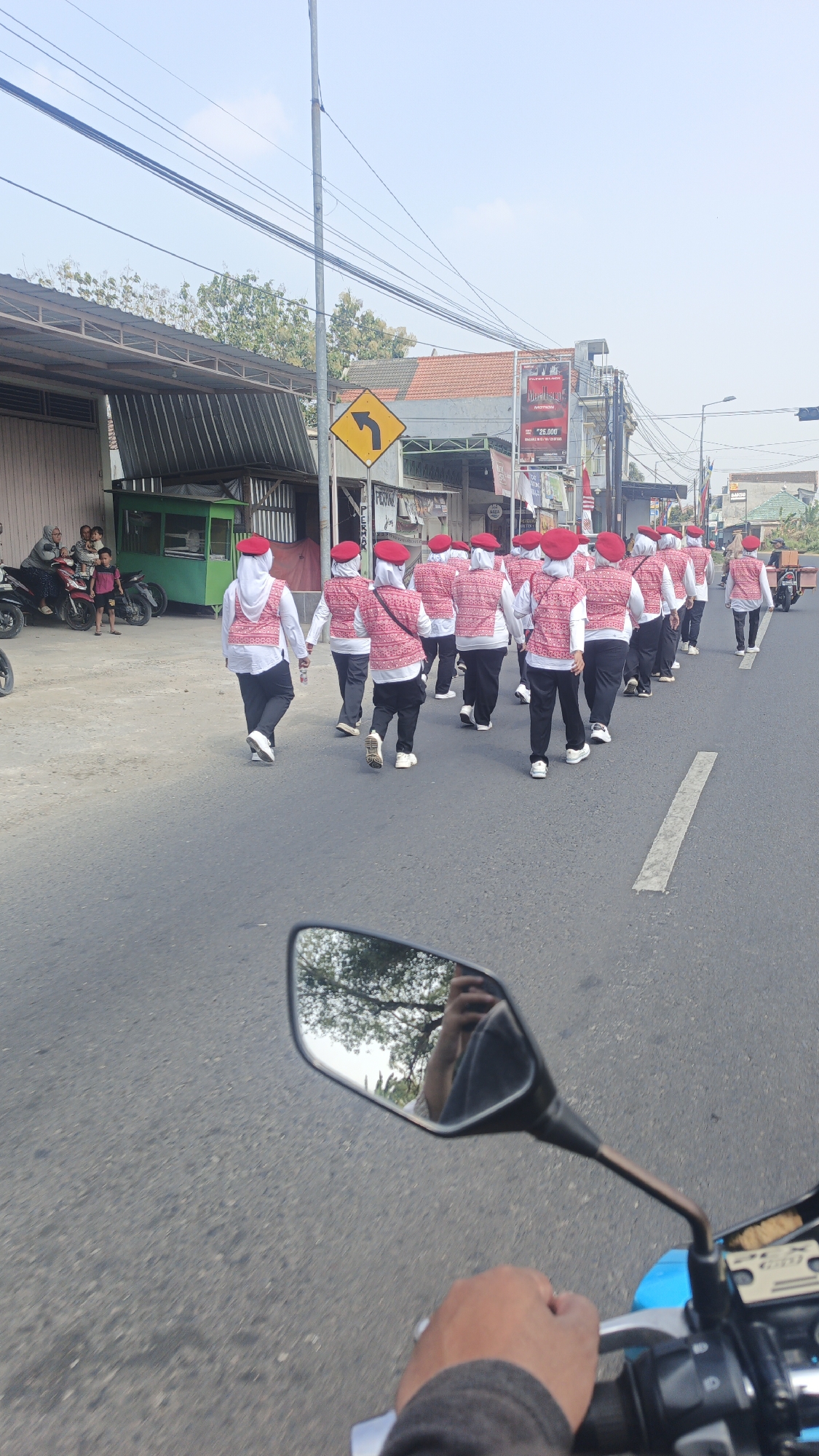 In Mlaten, Indonesia, a group of individuals wearing matching red berets and patterned vests walk in a parade down a paved road. The group, consisting of both adults and children, is walking in the same direction, on the right side of the road. The background shows various buildings and businesses, including a green stall or shop, and a billboard. The sky is clear and blue. A motorcycle rider's hand and a rearview mirror are visible in the foreground, suggesting the photo was taken from a moving motorcycle.