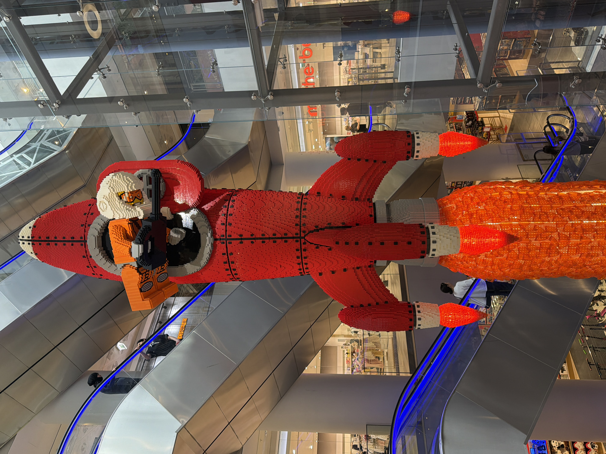 The image displays a large, red, brick-built rocket ship, likely made of Lego, suspended in the center of a multi-story building. The rocket is depicted with a Lego astronaut figure in the cockpit, wearing an orange spacesuit and a white helmet with orange goggles. The rocket's tail features an orange, fiery exhaust, also constructed from bricks.
The setting appears to be a modern indoor space, possibly a shopping mall or a public atrium, with escalators visible on either side. These escalators have blue lighting strips along their edges. Several people are seen on the escalators, dressed in casual clothing. Reflections are visible on the glass panels and metal structures of the building, showing more of the interior and suggesting a brightly lit environment.
The overall scene suggests an exhibition or a display, showcasing the intricate brick construction of the rocket. There is no indication of the time of day or weather conditions, and no discernible text is present in the image. The focus is on the impressive Lego creation and its installation within the architectural space.