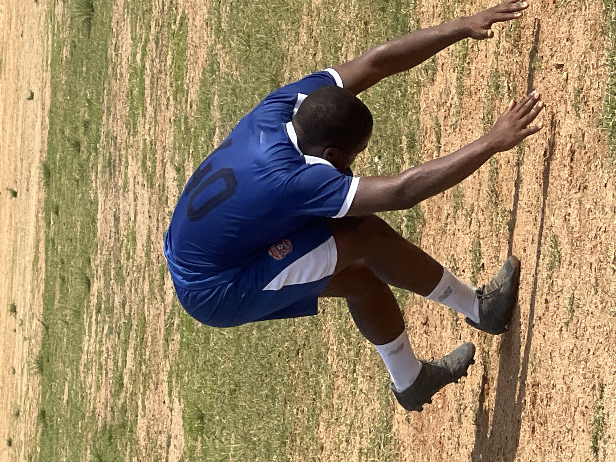 A person wearing a blue soccer uniform with the number 10 on the back and white trim is on a dry, grassy field. The person is in a bent-over, athletic stance, with their hands on the ground and their legs positioned as if preparing to spring forward. The uniform includes blue shorts with white side panels and a red and white crest on the left thigh. White soccer socks are visible, and the person is wearing dark soccer cleats. The ground is a mix of dry dirt and sparse, yellowish-green grass. The background shows a similar dry, open field with some patches of green vegetation. The lighting suggests it is daytime, possibly a sunny day, with shadows cast on the ground. The image appears to have been taken outdoors in an open area, characteristic of many public fields in Jos, Nigeria, where such sporting activities are common.