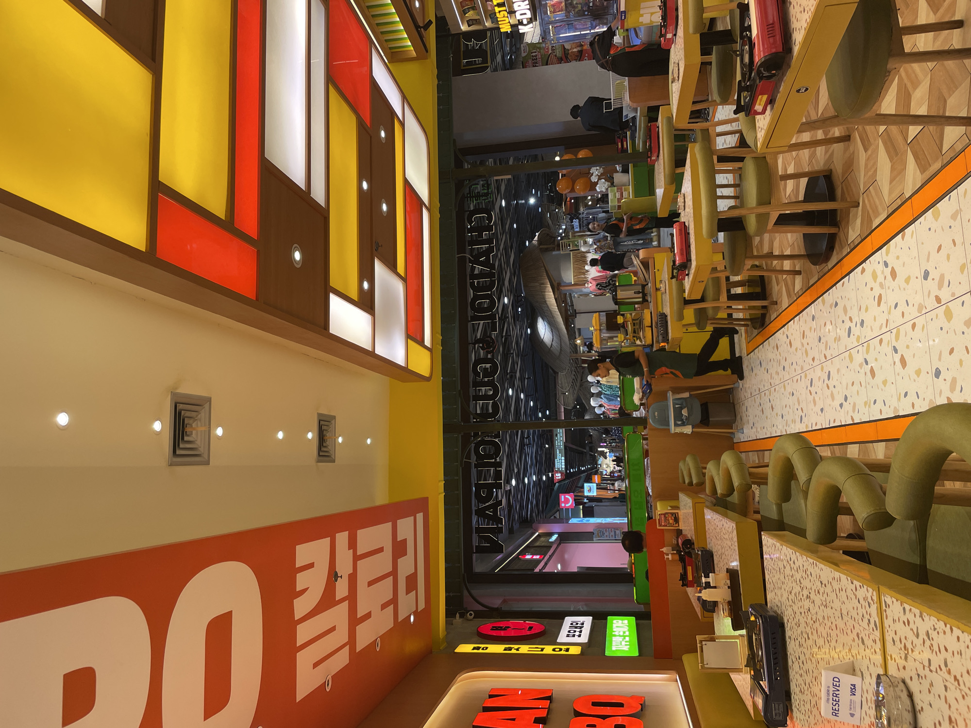 This image depicts the interior of a Korean barbecue restaurant, likely in Denpasar, Indonesia, given the presence of Korean signage and the general ambiance of a busy Asian mall or dining complex. The restaurant features vibrant yellow and orange decor, with large text on the ceiling and walls, possibly indicating the restaurant's name or theme. Tables are set with individual portable grills and condiments, indicating a self-cook barbecue experience. Several customers are visible, some seated at tables, while others appear to be staff members attending to diners or preparing food. The overall environment is brightly lit and appears to be a commercial space with a lively atmosphere, possibly a food court or a section of a shopping mall. The floor has a distinctive patterned tile design, and the ceiling incorporates a colorful, geometric lighting structure. A sign on one table indicates it is reserved and displays payment options like Visa.