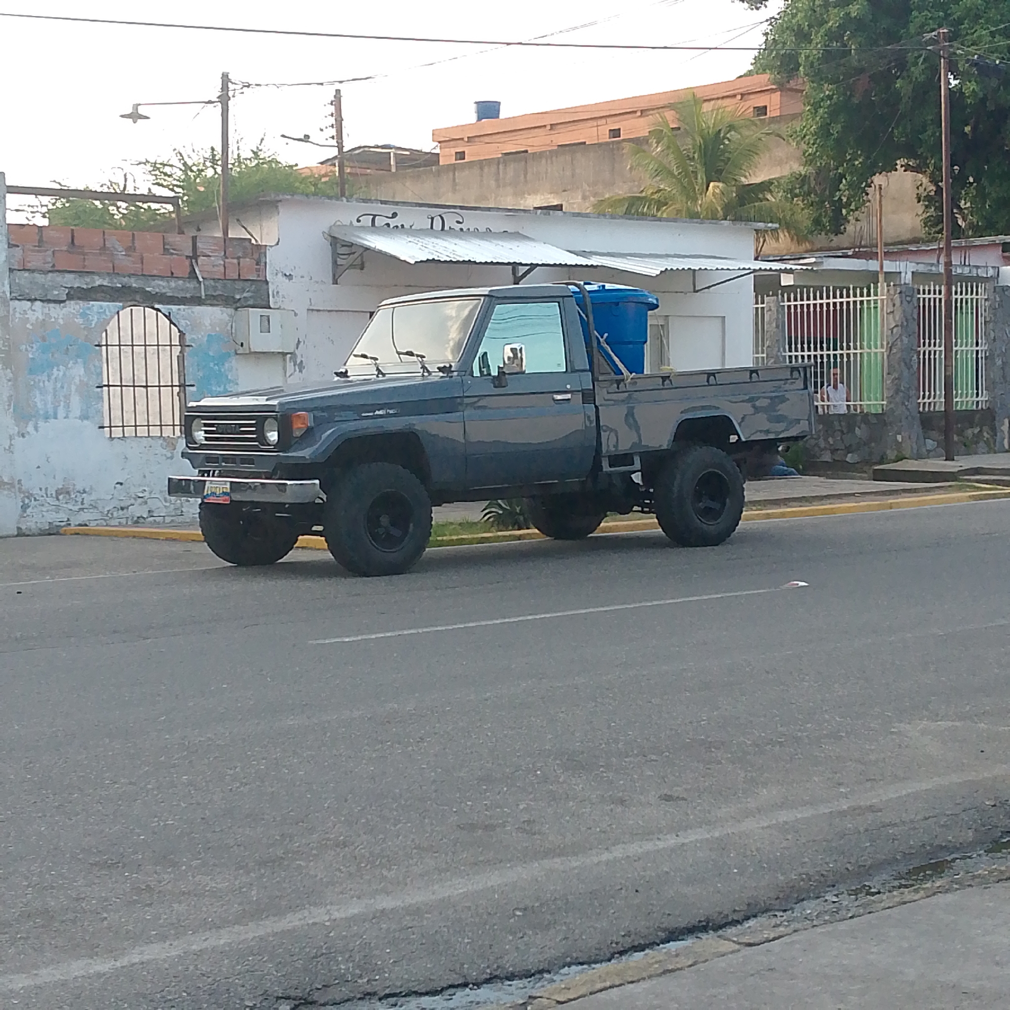 A dark gray, modified Toyota Land Cruiser 70 series pickup truck is parked on the side of a paved road in Tinaquillo, Venezuela. The truck features large, black-rimmed off-road tires and a prominent front bumper. A blue plastic barrel or water tank is secured in the bed of the truck. "TOYOTA" is visible on its grille, and a license plate with Venezuelan flag colors displays "30M15D".
In the background, a light-colored building with a weathered, textured wall and an arched, barred window is seen to the left. Above the truck, an awning made of corrugated metal extends from another white building. Partial text, "Tina D...", is visible on the wall above the awning, possibly indicating a business name. Further back, another building with an orange-brown facade and a blue water tank on its roof is visible.
To the right of the truck, there's a white metal gate with colorful panels (green and red) behind a stone wall. A person in a white shirt is partially visible sitting behind this gate. Dense green foliage, including palm trees, and utility poles with overhead lines are scattered across the background. The sky is bright and clear, suggesting daytime, likely late afternoon or early morning due to the soft light. A streetlamp on a utility pole is visible in the upper left. Another person appears partially obscured near the truck's rear wheels, sitting on the ground behind the fence. The road surface is asphalt, with a yellow curb line visible on the right.