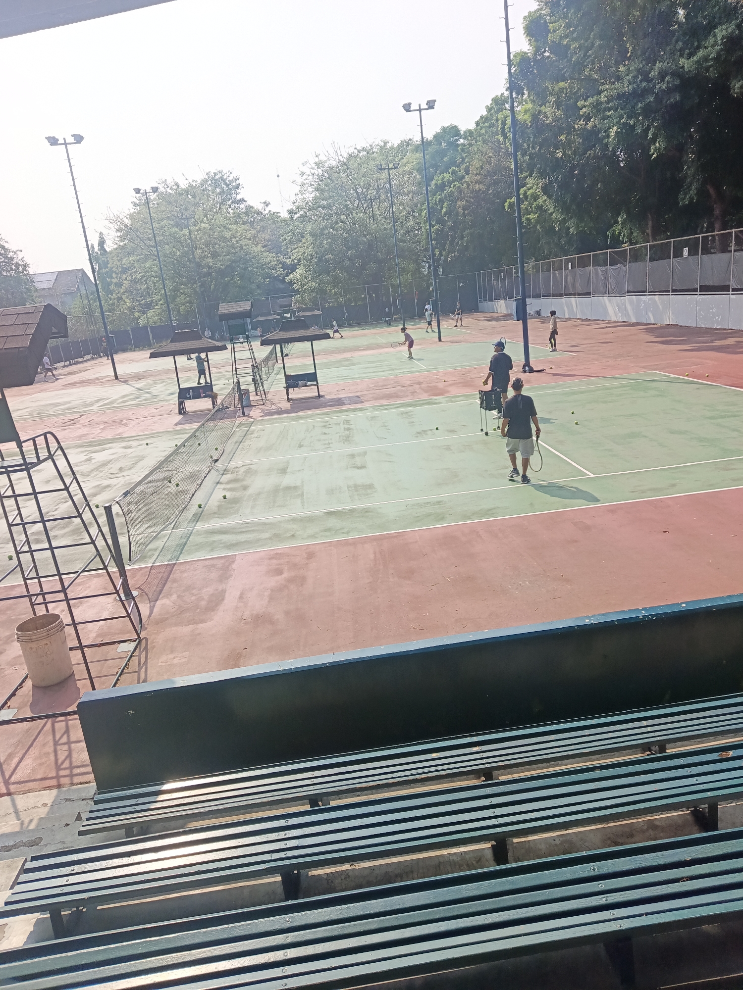 The image depicts a tennis court, likely located in East Jakarta, Indonesia, on a bright, sunny day. The court surface is a mix of reddish-brown and green, with visible court lines. Several people are present, engaged in tennis-related activities.
In the foreground, a set of empty green bleachers is visible, partially in shadow. To the left, a white bucket and a metal ladder leading to a umpire's chair are present.
Further onto the court, several tennis nets are set up. Some people are actively playing or practicing tennis. One individual is seen from behind, holding a tennis racket and facing away from the camera. Another person is standing near a basket of tennis balls, possibly serving as a coach. Other figures are scattered across the court, some in motion, suggesting a training session or a game in progress. Tennis balls are visible on the court surface.
The background shows trees and foliage, consistent with an outdoor sports facility. Light poles with multiple lamps are also present around the court, indicating the possibility of evening play. The overall scene is one of a lively sports environment under clear skies.