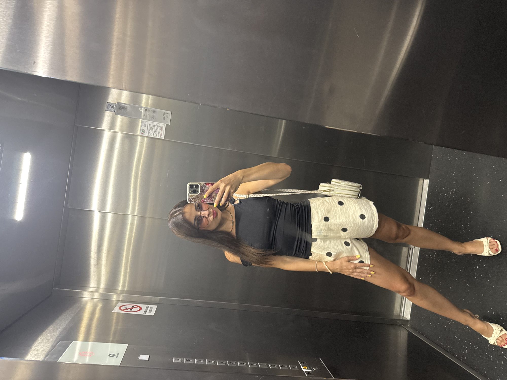 A woman is taking a mirror selfie in an elevator. She is wearing a black tank top and a white skirt with black polka dots. She has on white sandals and a white purse is slung over her shoulder. She is wearing sunglasses and has her hair down. She is holding her phone up to take the selfie. The elevator has stainless steel walls and a black floor. There are some signs on the wall of the elevator. One sign has a red circle with a line through a person, indicating no smoking. Another sign is a safety notice. There is also an elevator panel with buttons. The lighting in the elevator is bright. The overall impression is that the woman is dressed stylishly and is confident.
