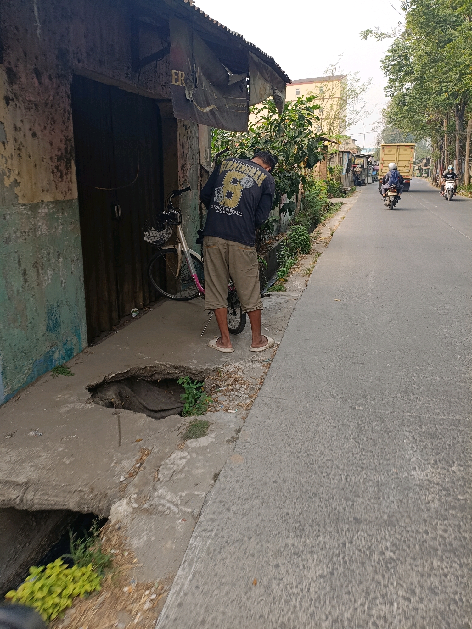 A man is standing with his back to the camera, facing a crumbling section of sidewalk next to a building in Tangerang, Indonesia. He is wearing a black long-sleeved shirt with yellow lettering and tan shorts. A bicycle with a basket is parked next to him. The sidewalk has a large hole in it, with plants growing out of the darkness below. The street is paved and lined with trees. A yellow truck and two motorcycles are visible further down the street. The building appears to be old and weathered, with peeling paint and a corrugated metal roof. The sky is overcast.