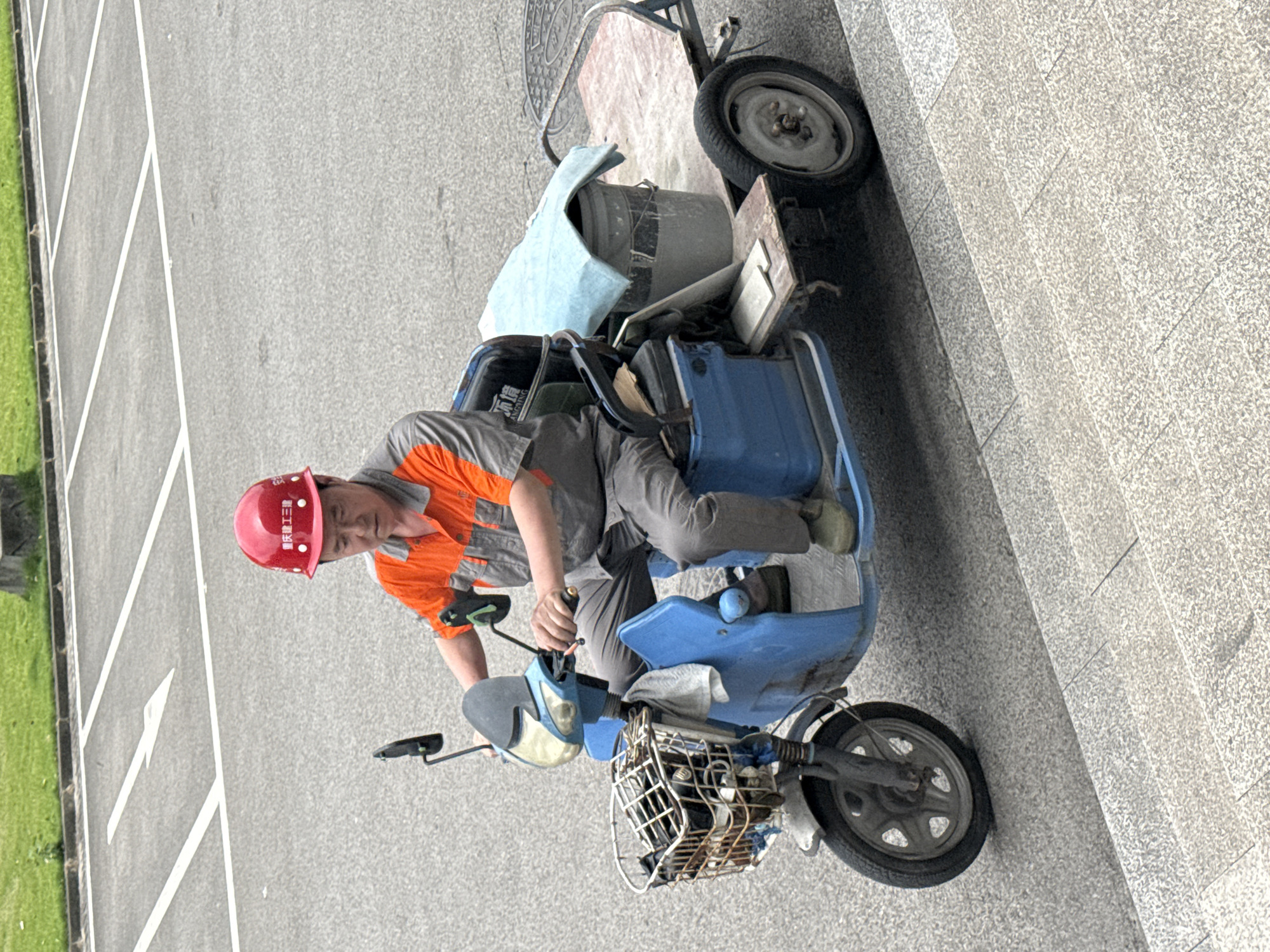 A worker with a red helmet is riding a blue electric tricycle in Jiangjin, China. The tricycle is equipped with a front basket, and a cargo rack in the back carries various items including a bucket. The worker is wearing an orange and gray uniform. He is traveling on a paved road alongside a grassy area. The scene is well-lit.