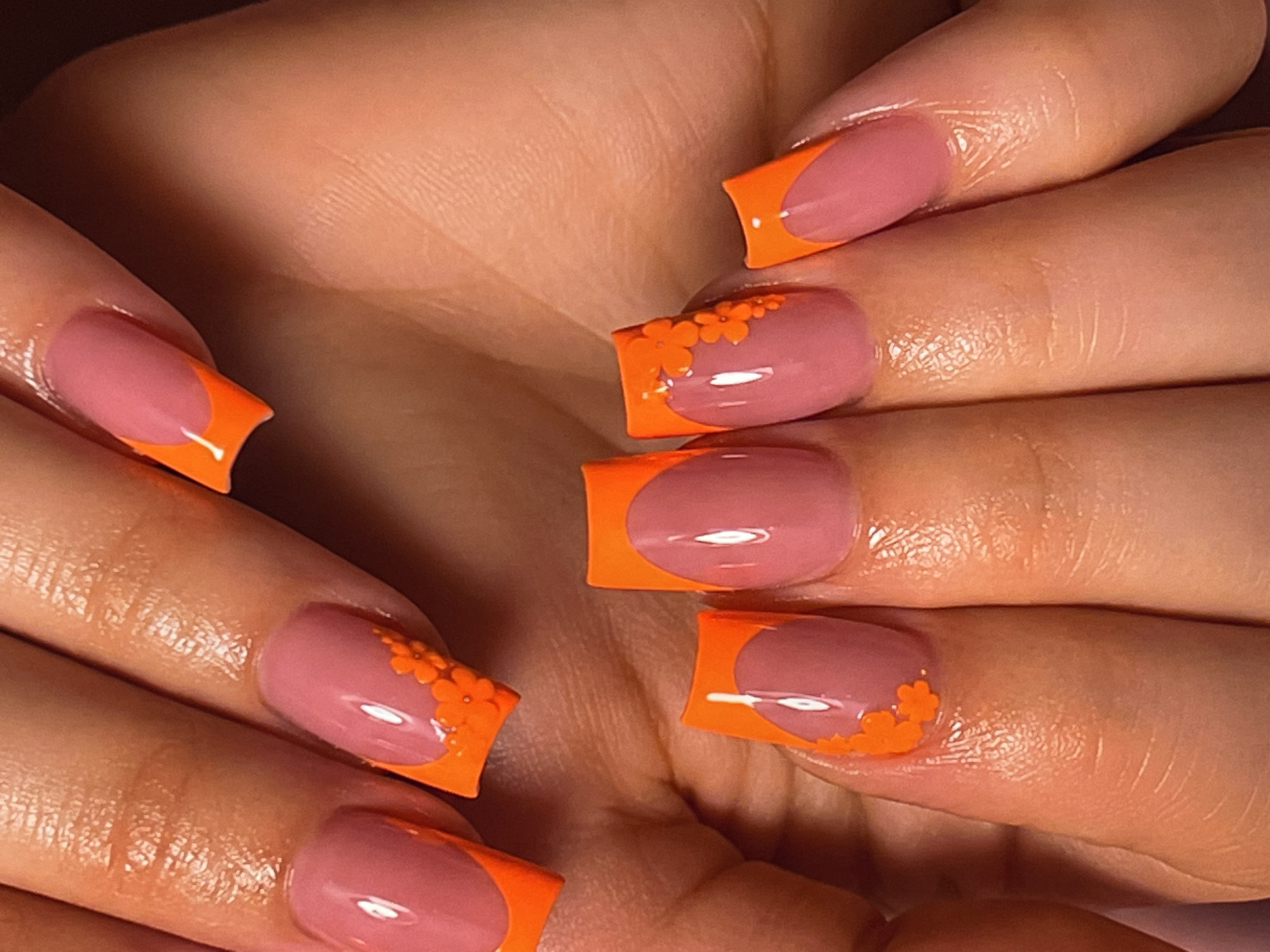 This close-up media file shows a person's hands with a French manicure. The nails are a medium length and have a square-rounded shape. The base color of the nails is a soft pink, and the tips are painted in a vibrant orange. Some of the nails are decorated with small, bright orange floral designs placed along the edge of the orange tip. The skin of the hands appears well-moisturized, with a slight sheen. The background is out of focus and shows more of the person's hands and fingers, suggesting the photo was taken from the perspective of someone looking at their own hands. There is no visible text in the image. The lighting is warm, and the overall impression is one of attention to detail and personal grooming. The activity taking place is simply the presentation of the manicure. The emotion conveyed is one of polished appearance and possibly pride in the nail art.