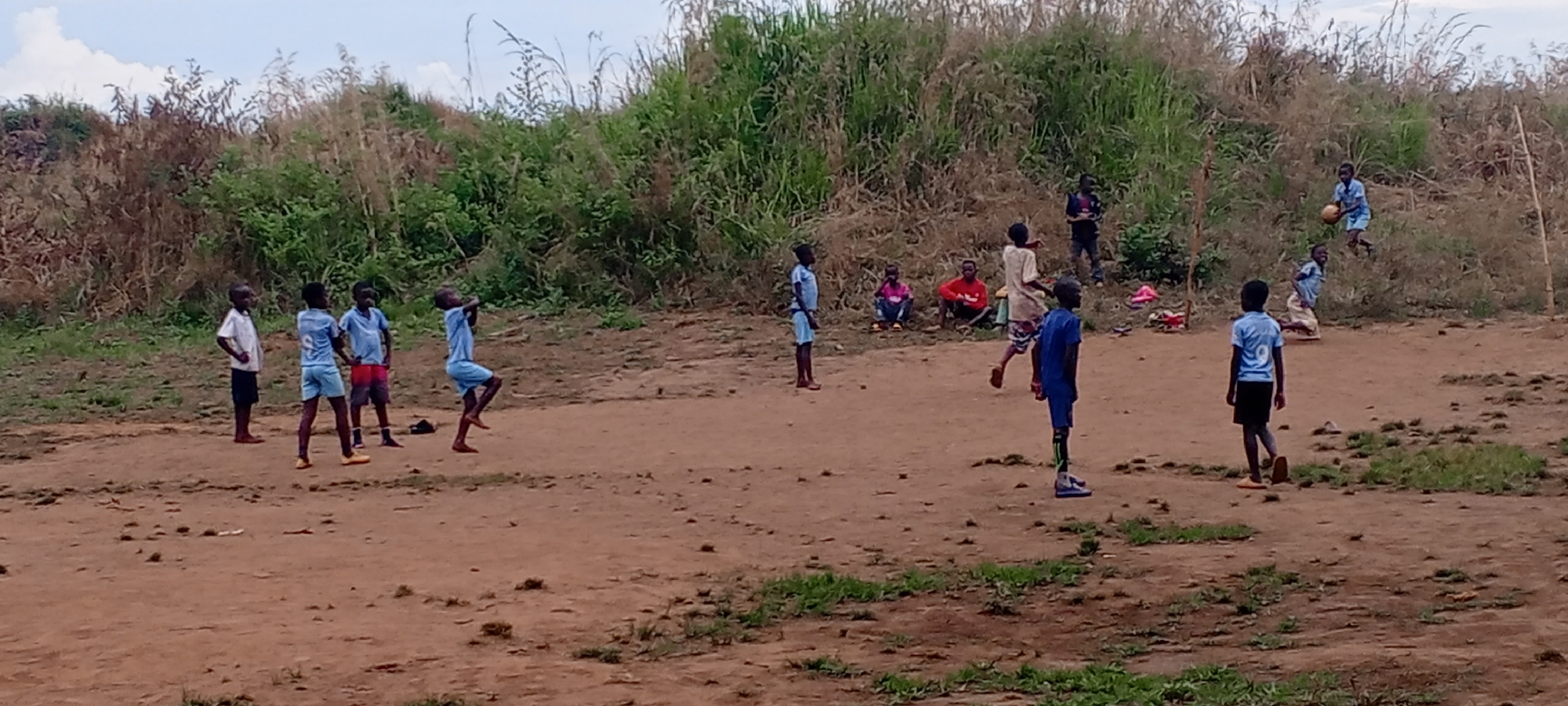 The image depicts an outdoor scene in Ngalula, Democratic Republic of the Congo, where a group of approximately a dozen children and young people are engaged in an informal game of soccer. The setting is a reddish-brown dirt field with uneven terrain and scattered patches of green grass. A dense, overgrown bank of tall green and dry brown vegetation forms the background, suggesting a rural or semi-rural environment. The sky is overcast, indicating diffused daylight, likely late morning or afternoon.
The primary activity is a soccer game. On the left side of the frame, several boys in light blue jerseys and dark shorts are visible, one appearing to jump for the ball, while another stands ready. Further right, a boy in a light blue jersey is holding a soccer ball near a slight incline. Another boy, viewed from behind, wears a light blue jersey with the number "9" clearly visible on his back. Two thin wooden poles are present on the right side of the field, likely serving as makeshift goalposts.
Beyond the active players, several individuals are observing or resting. On the far left, a boy in a white shirt and dark shorts stands. In the middle-background, three people are seated on the ground, two in red shirts and one in a pink shirt, seemingly spectators. Another person stands behind them in dark clothing. The overall scene conveys a sense of communal and casual sport, with children actively participating and others watching. No explicit emotional expressions are clearly discernible due to distance, but the atmosphere appears relaxed and engaged.