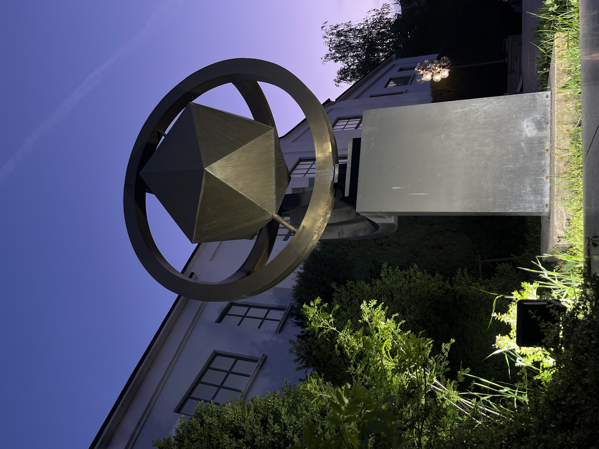 The image captures an outdoor scene at dusk or night in Sofia, Bulgaria, featuring a large, metallic geometric sculpture as its central subject. The sky above is a deep indigo or purple, with a faint contrail visible.
The sculpture consists of a substantial outer ring, within which an elaborate, multifaceted polyhedron is suspended. The polyhedron appears to be a stellated octahedron or a similar complex geometric shape, crafted from a dark, possibly bronze or brass, metal that reflects some light. The entire sculpture is presented from a low-angle perspective, emphasizing its size and intricate form against the evening sky.
In the background, a light-colored building with multiple rectangular windows and a sloped roof is visible. The building's architecture suggests a European style. Further back and to the right, a street lamp with a cluster of spherical lights, reminiscent of grapes, illuminates the area.
The foreground and midground are dominated by lush green vegetation, including various bushes and trees. Some of these plants are brightly illuminated from below by unseen spotlights, creating strong contrasts between light and shadow. To the right of the sculpture, a dark, rectangular, flat panel or sign stands upright, next to what appears to be a concrete or wooden block structure partially covered by grass. The overall lighting scheme, combining artificial light with the twilight sky, creates a dramatic and serene atmosphere. No discernible text is visible anywhere in the image.