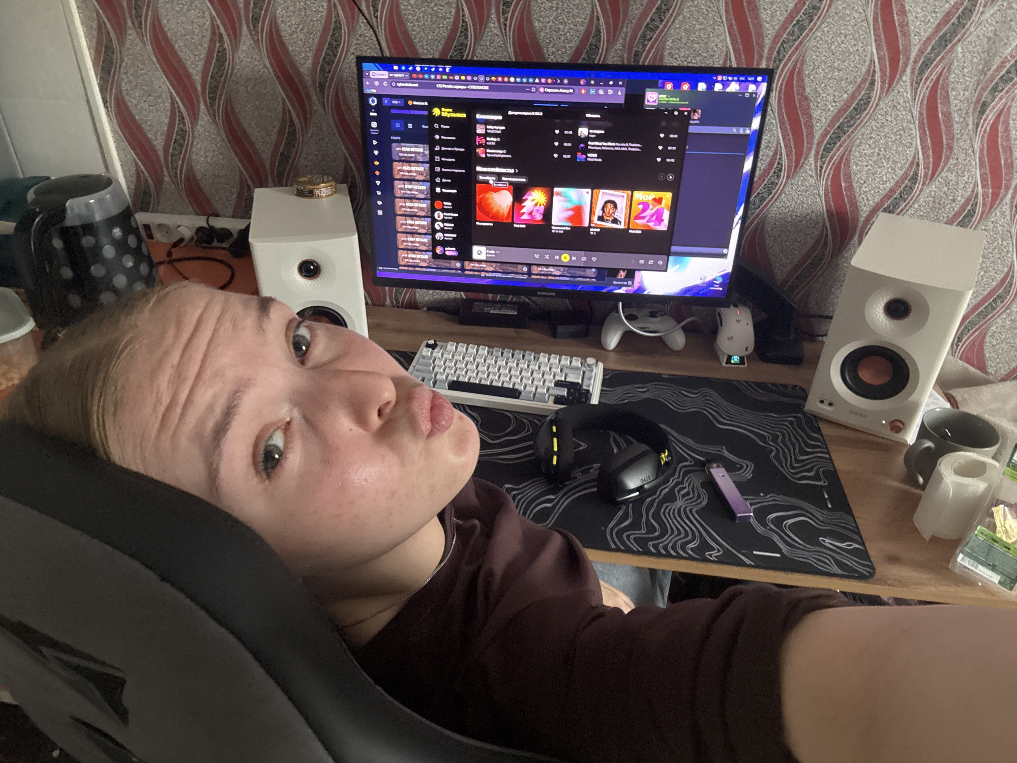 The image shows a woman sitting in a gaming chair, facing the camera, in a room with a computer setup. The woman has fair skin, blonde hair, and is wearing a brown long-sleeved shirt. Her expression is playful with pursed lips.
Behind her is a computer desk with a monitor displaying what appears to be a music streaming service, possibly Spotify, with a playlist visible on the screen. On the desk are white computer speakers on either side of the monitor, a white mechanical keyboard, black and yellow headphones, a large black mousepad with a topographic pattern, and a purple vaping device. There is also a mug, a cylindrical object (possibly a candle), and some small packaged items near the right speaker.
The wall behind the desk is covered with wallpaper featuring a wavy pattern in shades of brown and red. There is also a black kettle and a white cabinet visible to the left of the desk.
While the city of Perm, Russia, is not definitively discernible from the image, the style of the interior and the items present suggest a possible location in a residential area, possibly in Russia.