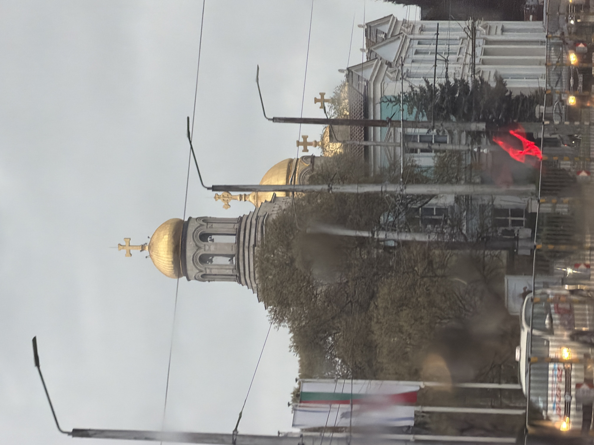 The image displays an urban scene dominated by a large Orthodox Christian church featuring a prominent bell tower and multiple golden onion domes, each topped with a cross. The church, with its ornate architectural details, is partially obscured by trees, some with bare branches, suggesting a colder season or early spring.
The sky is uniformly grey, indicative of an overcast day. Numerous utility poles and wires crisscross the upper portion of the frame, hinting at urban infrastructure like trolleybus lines. In the lower left foreground, a Bulgarian flag, distinguishable by its horizontal white, green, and red stripes, is visible, flying from a pole.
On the right side of the image, a large vehicle, possibly a bus or trolleybus, is partially visible, with reflections and lights suggesting it is in motion or parked on a street. Other buildings can be seen in the background to the right of the church. A blurry, vivid red shape is present in the mid-right, which could be a reflection or a light source. The overall image quality suggests it was taken through a wet or smudged window, possibly during or after rain. The scene captures a typical city environment centered around a significant religious landmark in Varna, Bulgaria.