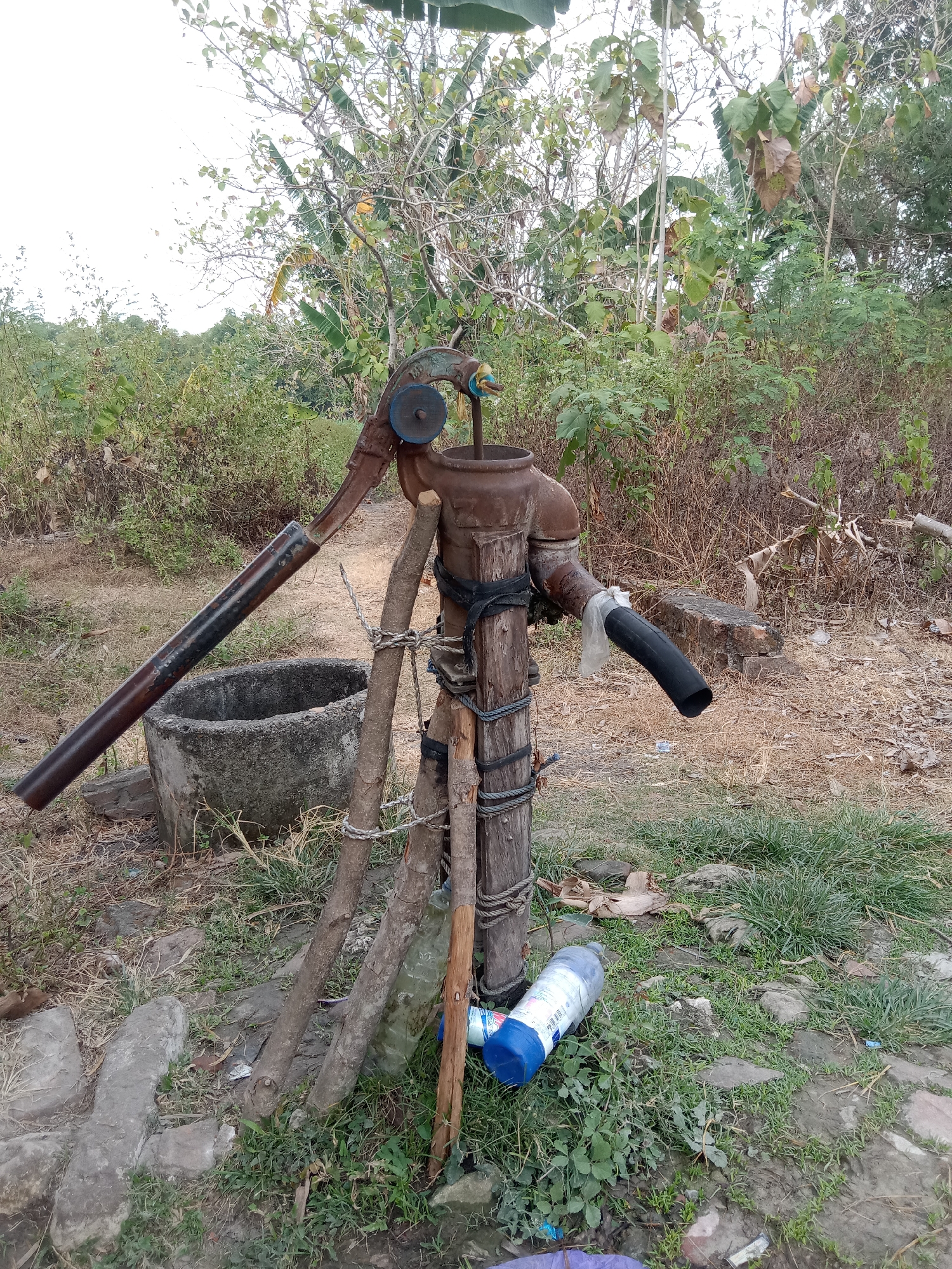 An outdoor scene in Jumput, Indonesia, features an old, rusty hand pump for well water. The pump is made of metal and is mounted on a wooden post secured with rope. A concrete basin sits to the left of the pump, and a black hose is attached to an outlet pipe, directing water away from the pump. Several pieces of wood are propped up around the pump, and a plastic bottle lies on the ground nearby. The background consists of dense vegetation and trees under a cloudy sky.