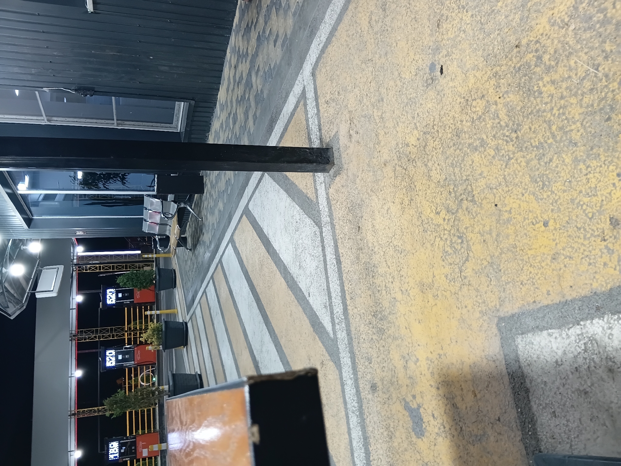 The image depicts an outdoor scene at night, likely a gas station, given the presence of gas pumps with illuminated displays. There are at least three gas pumps visible, two with the number "520" displayed and one with "189". The pumps are situated under a covered structure with lighting. Several potted plants are placed in front of the pumps. In the background, there's a building with large windows, and some seating arrangements, possibly benches, are visible. The ground is paved with distinct yellow and white markings, resembling a crosswalk or designated area. A dark, rectangular pillar is positioned in the foreground, partially obscuring the view. The overall lighting suggests artificial illumination from the gas station and surrounding structures. The image does not provide any information or visual cues to suggest the location is in Sayat Nova, Armenia.