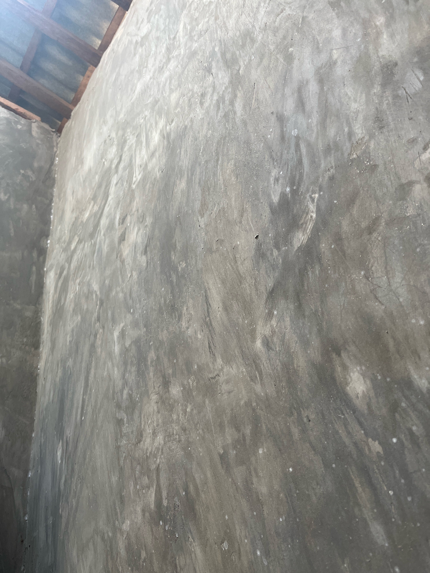 The image shows an interior corner of a structure, likely under construction, in Kebumen, Indonesia. The walls are bare concrete with visible trowel marks and variations in shading, suggesting a raw or unfinished aesthetic. In the upper left portion of the image, a portion of a roof structure is visible, consisting of wooden beams and corrugated metal sheeting. The lighting suggests it is daytime, with light entering from the roof area. There are no people or active construction processes depicted in the image. The overall impression is one of an unfinished building interior.
