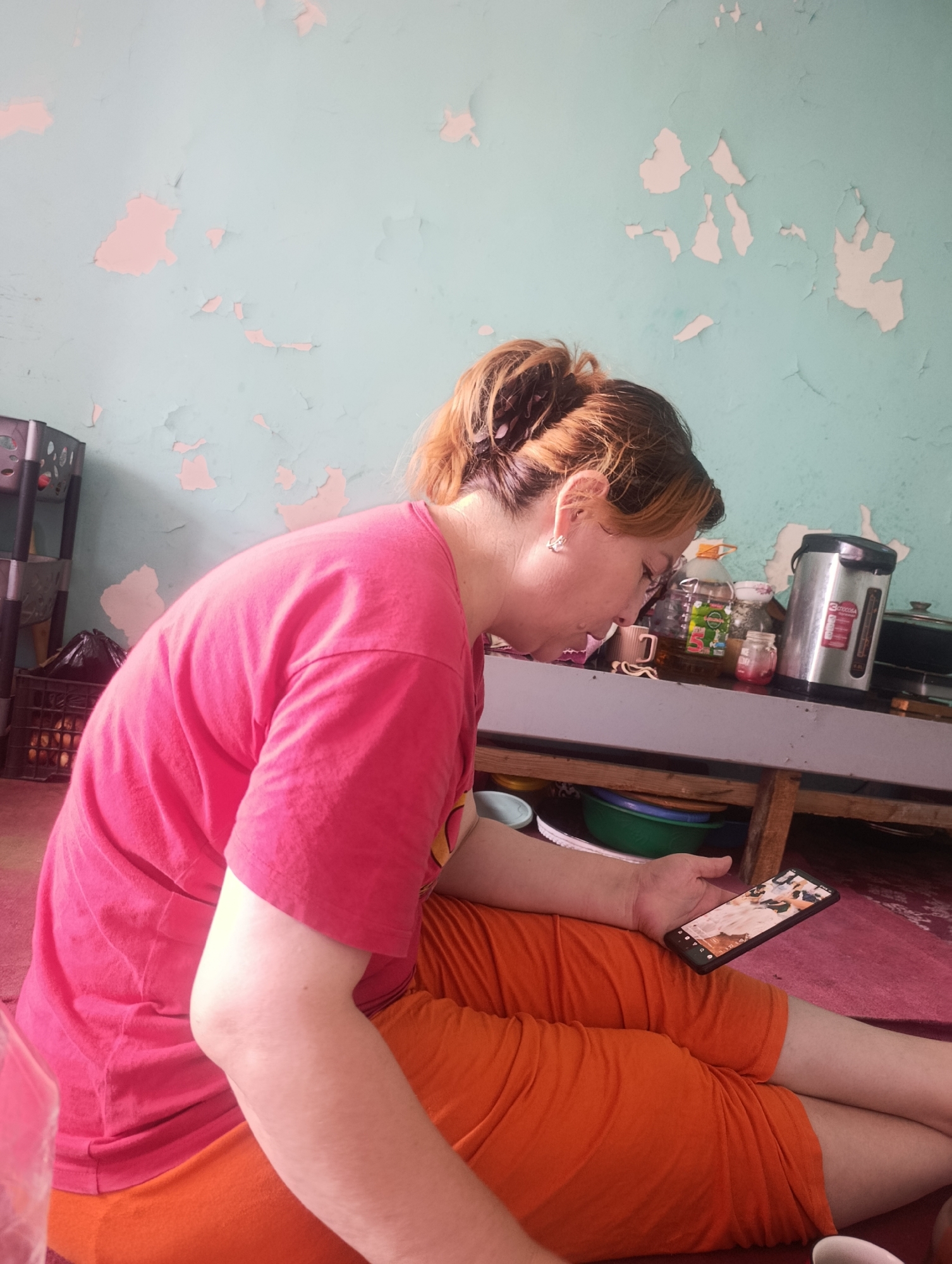 A woman with her hair pulled back in a bun, wearing a pink t-shirt and orange pants, is sitting on the floor and looking at her phone. She is in a room with a light blue wall that has peeling paint. There are shelves behind her with various items on them, including a silver electric kettle and a bottle of oil. The floor is covered with a red rug. The scene is likely a residential interior in Marhamat, Uzbekistan.