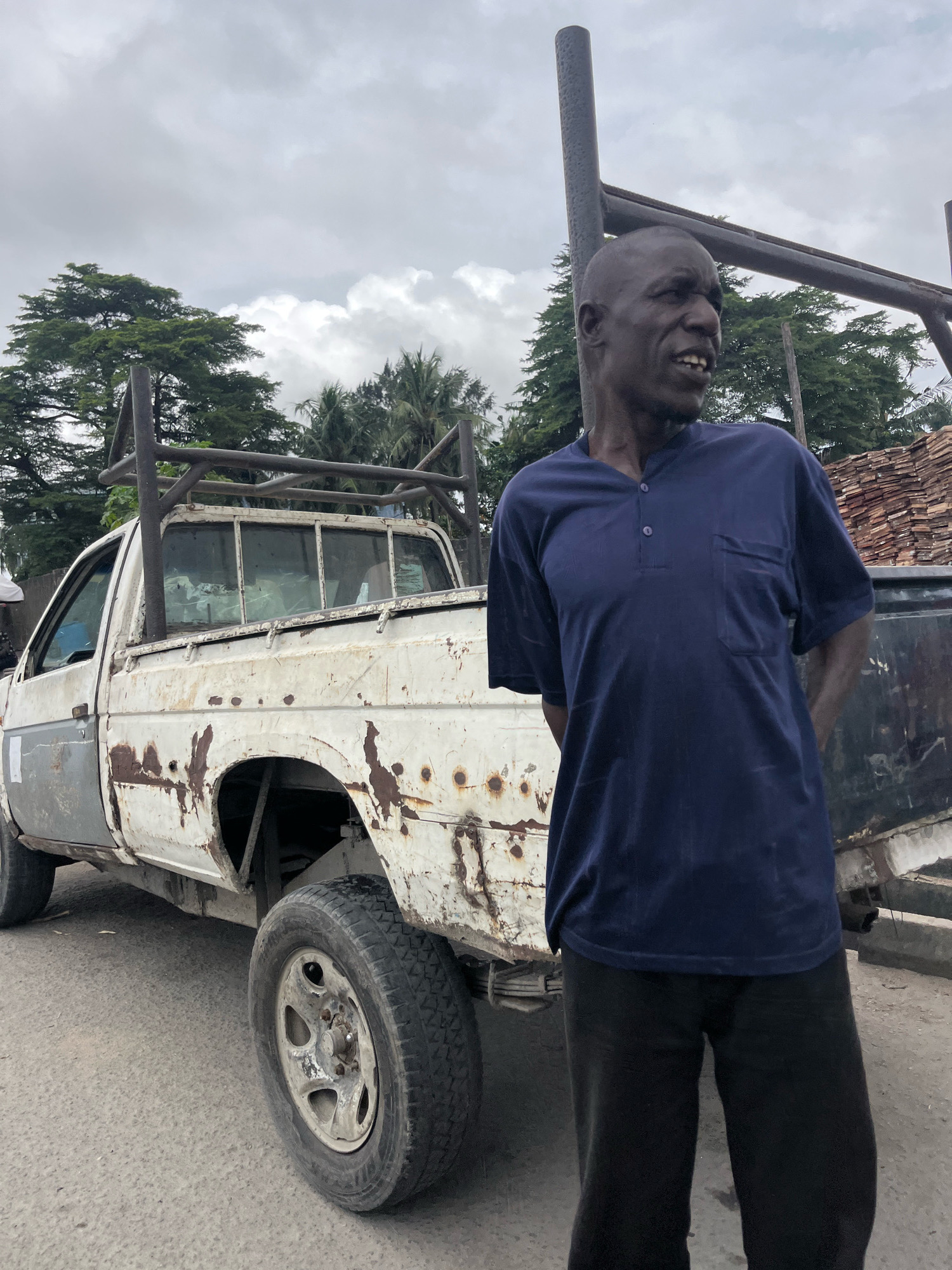An African man stands to the right of a heavily rusted, white pickup truck in Ado, Nigeria. The man, with a shaved head and a dark complexion, wears a navy blue t-shirt with a pocket and black pants. His hands are clasped behind his back as he looks to his left. The truck, which appears old and weathered, has a metal rack structure over its bed. Piles of wood or similar materials are stacked to the right of the truck. The background consists of trees and a cloudy sky. The ground is paved, suggesting an outdoor urban or semi-urban setting.