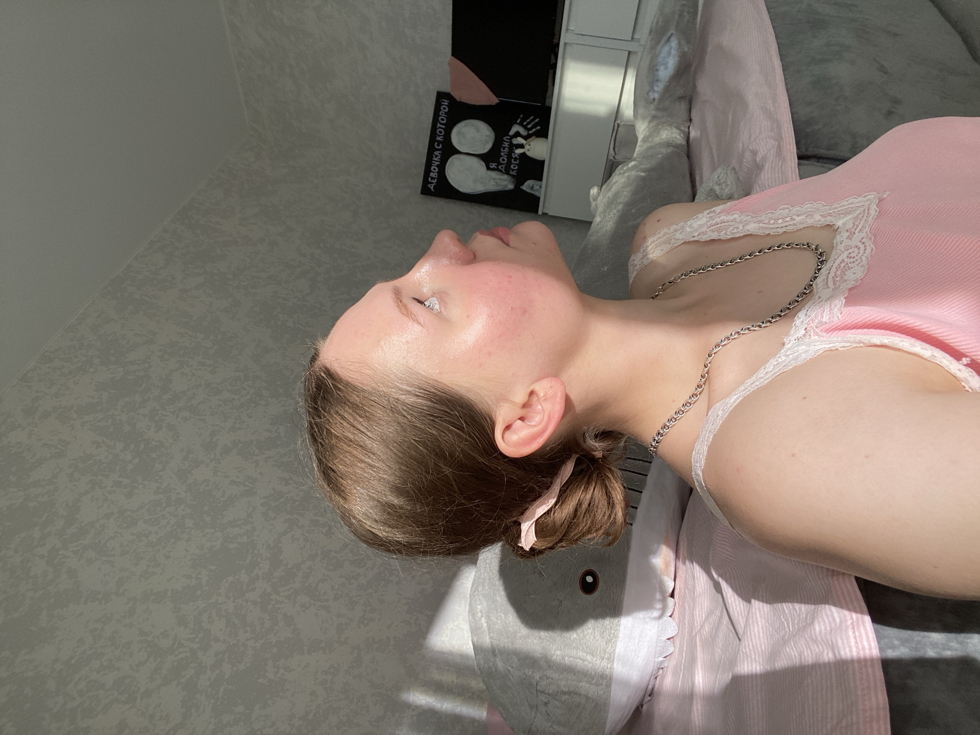 The media file shows a young woman lying down, possibly on a bed or sofa, in what appears to be an indoor setting. The sunlight casts strong shadows, suggesting it is daytime, possibly late afternoon due to the angle of the light. The woman is facing away from the camera, looking upwards and to the side. She has her hair tied back in a bun with a pink ribbon. She is wearing a pink camisole with white lace trim and a silver chain necklace.
To the left of the woman, on a dark surface, is a book or card with text in Russian. The visible text includes "ДЕВОЧКА С КОТОРОЙ" and "Я ДОБИЛ КОСМ". There are also illustrations of circles, possibly representing planets or moons, and a drawing of a hand with lines radiating from it. The overall mood is relaxed, with the woman's pose and the soft lighting contributing to a calm atmosphere. The setting appears to be a room with textured walls. A large plush toy, possibly a shark, is visible beneath the woman. The context suggests this photo was taken in Izhevsk, Russia.